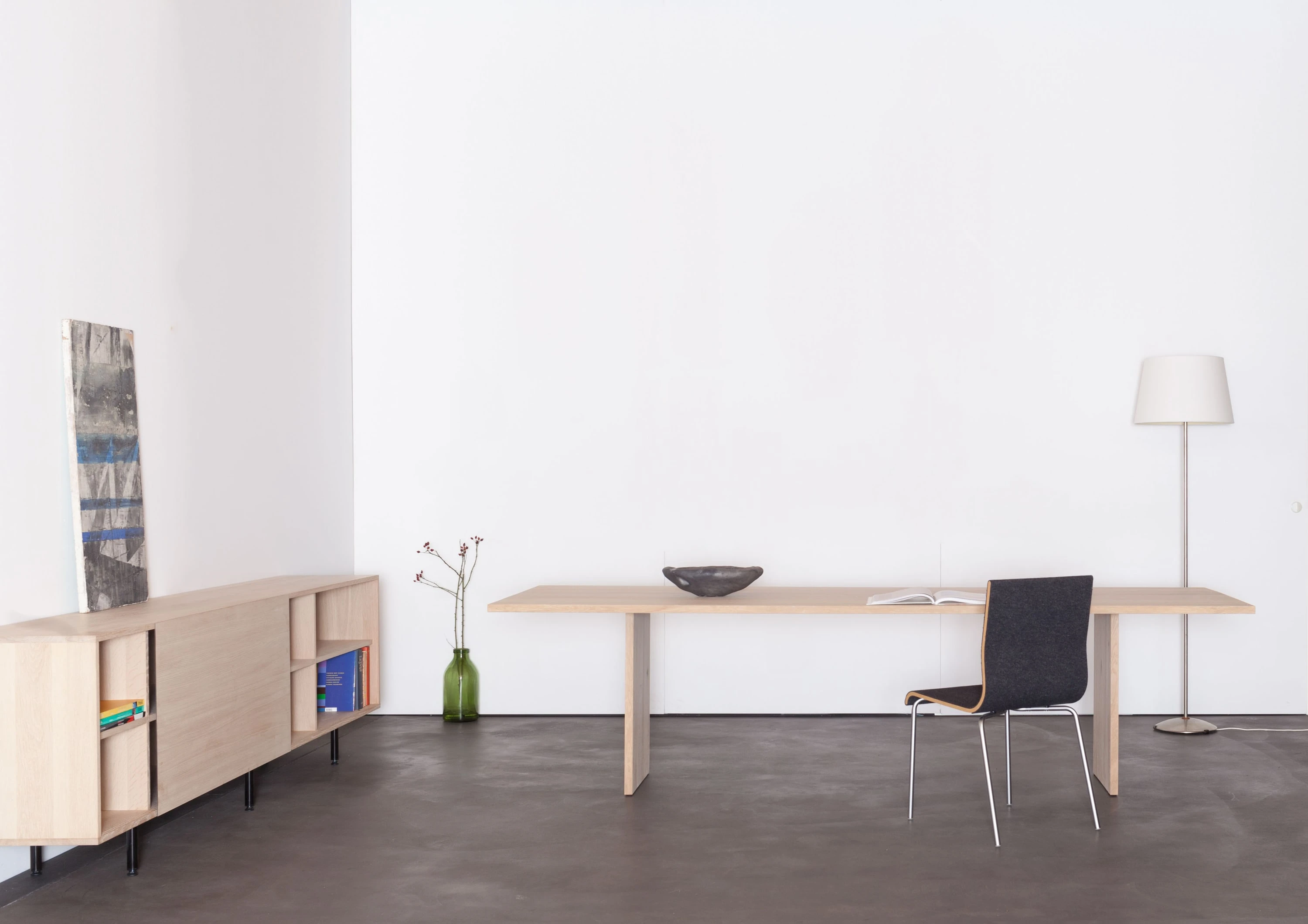 Arbeitsplatz mit schmalem Holztisch, schwarzem Stuhl und Stehleuchte, links ein niedriges Sideboard aus Holz mit Bild und Vase.