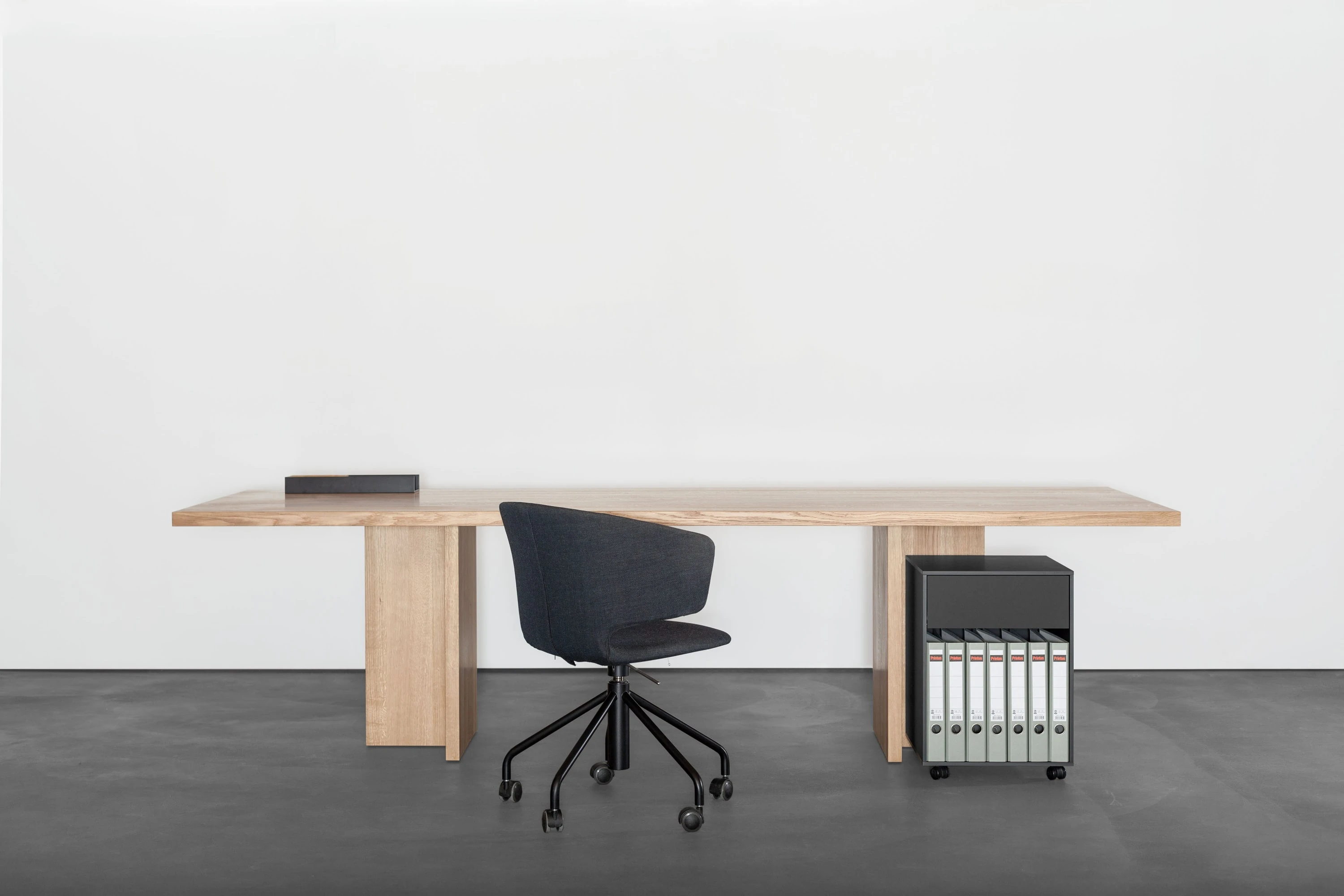 Schreibtisch aus Holz mit schwarzem Bürodrehstuhl und schwarzem Rollcontainer mit Ordnern.