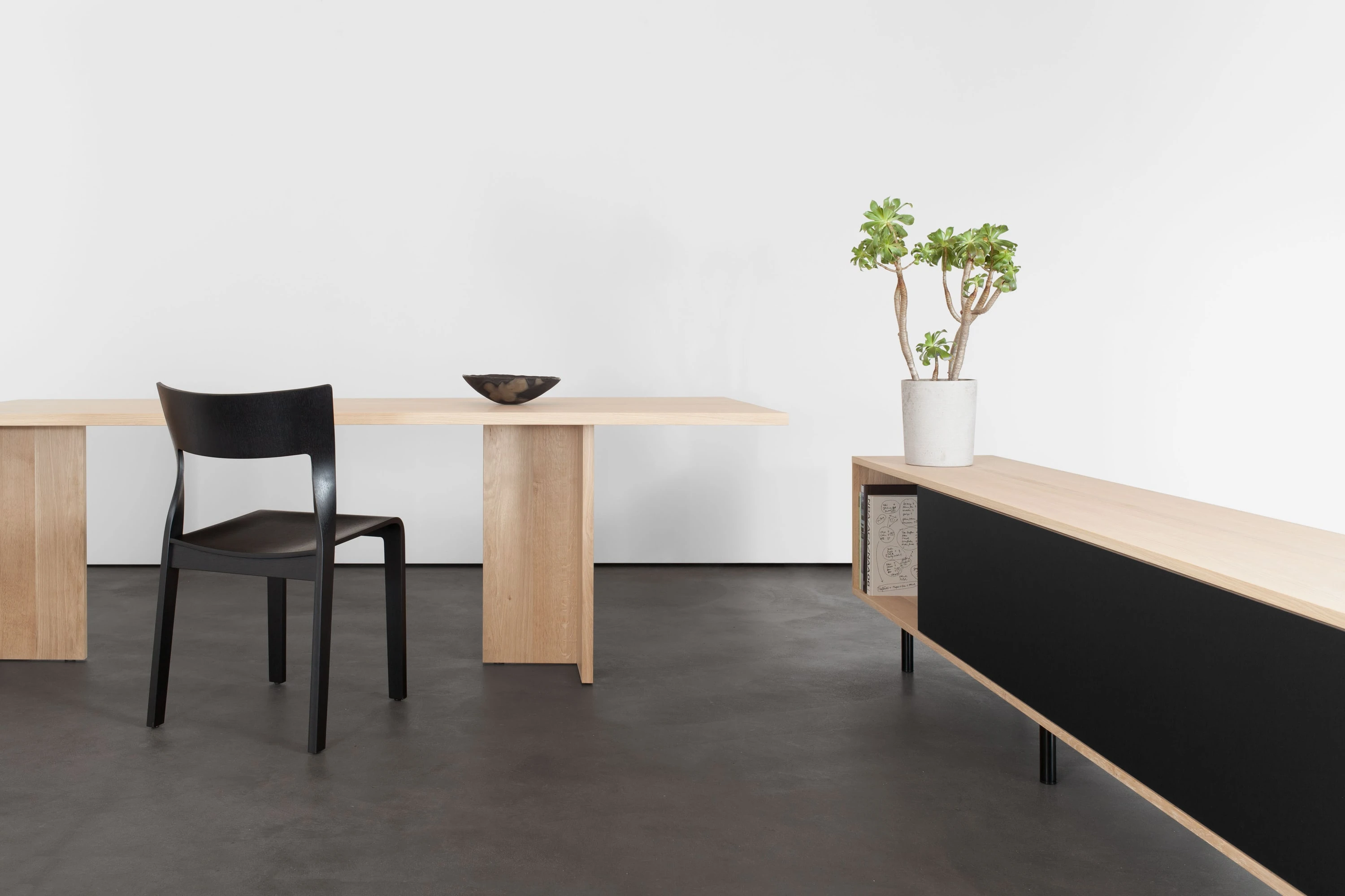 Klapptisch aus Eiche mit schwarzem Stuhl und Schale, rechts ein Sideboard mit schwarzer Front und Pflanze.