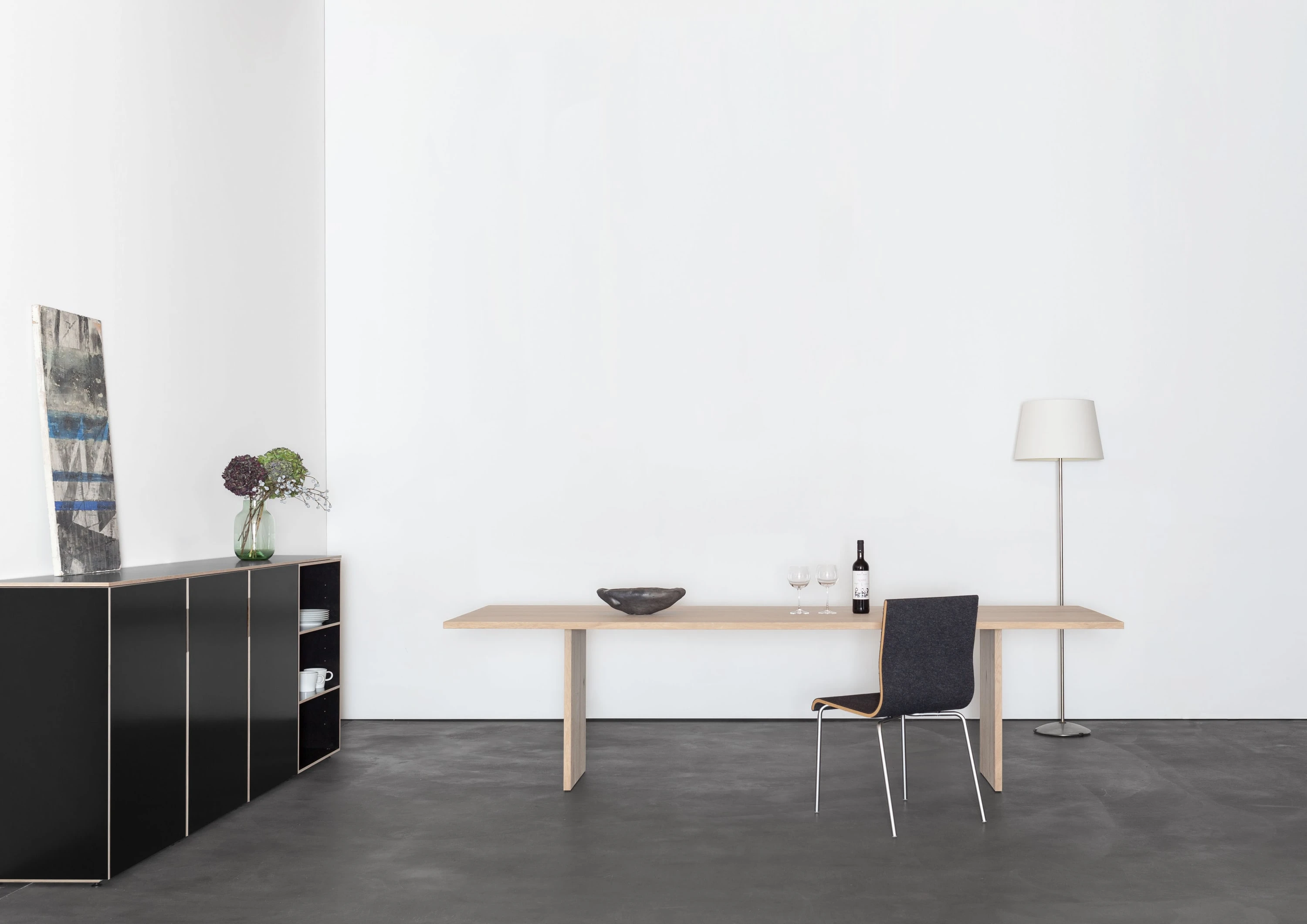 Schwarzes Highboard mit Bild und Blumen, davor ein Holztisch mit Weinflasche, Gläsern und grauem Stuhl, daneben eine Stehleuchte.