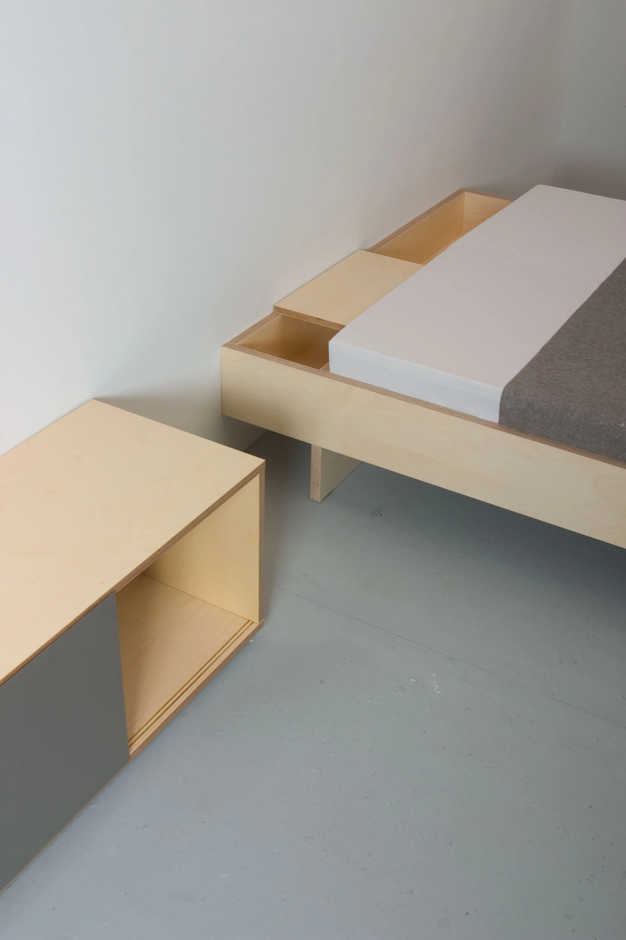 Ecke eines Bettes aus hellem Holz mit aufgeklapptem Fach, daneben ein niedriges Möbel mit dunkler Front.