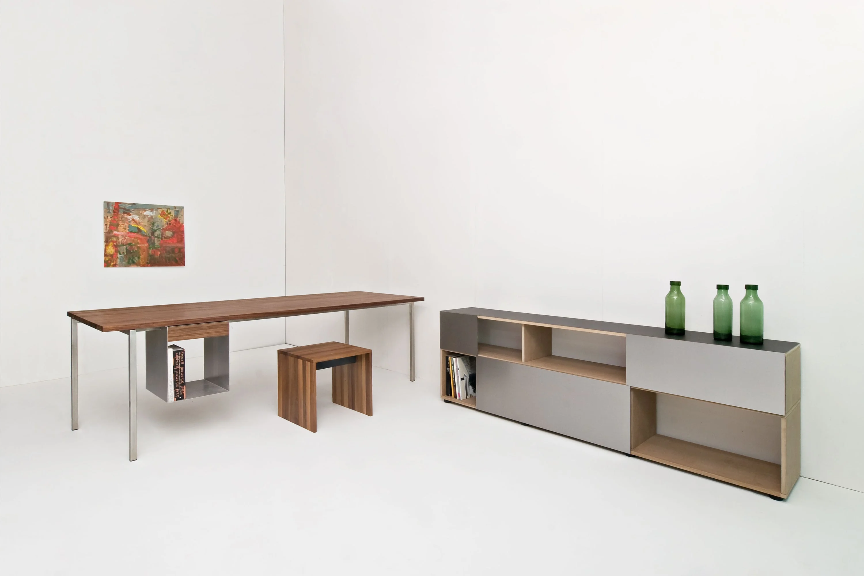 Tisch aus Nussbaum auf Stahlbeinen mit Hängefach und kleiner Bank darunter, rechts ein langes Sideboard mit grauen Fronten und grünen Flaschen.