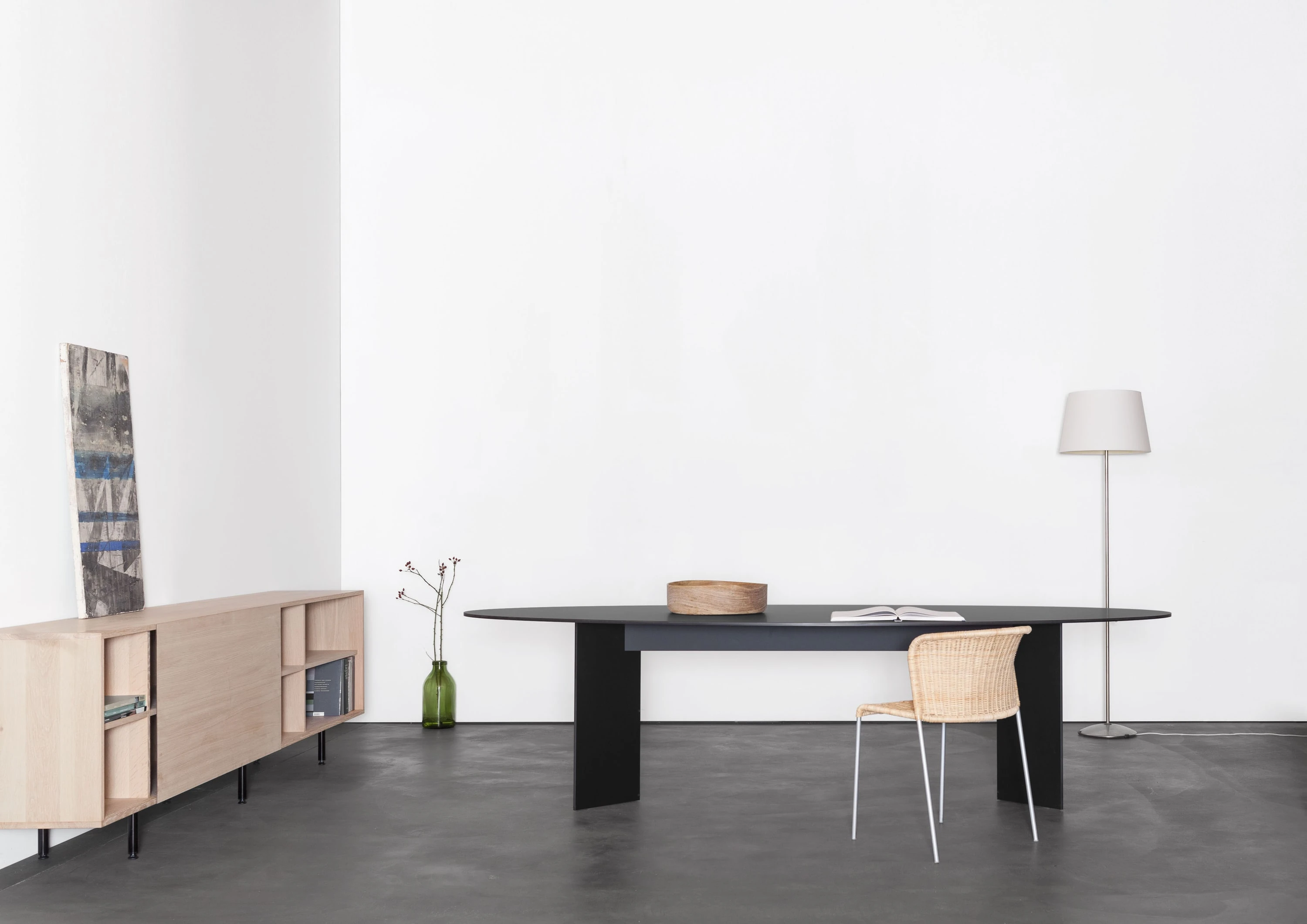 Langer schwarzer Tisch mit Geflechtstuhl, links ein Sideboard aus Eiche mit Bild und Vase, rechts eine Stehleuchte.