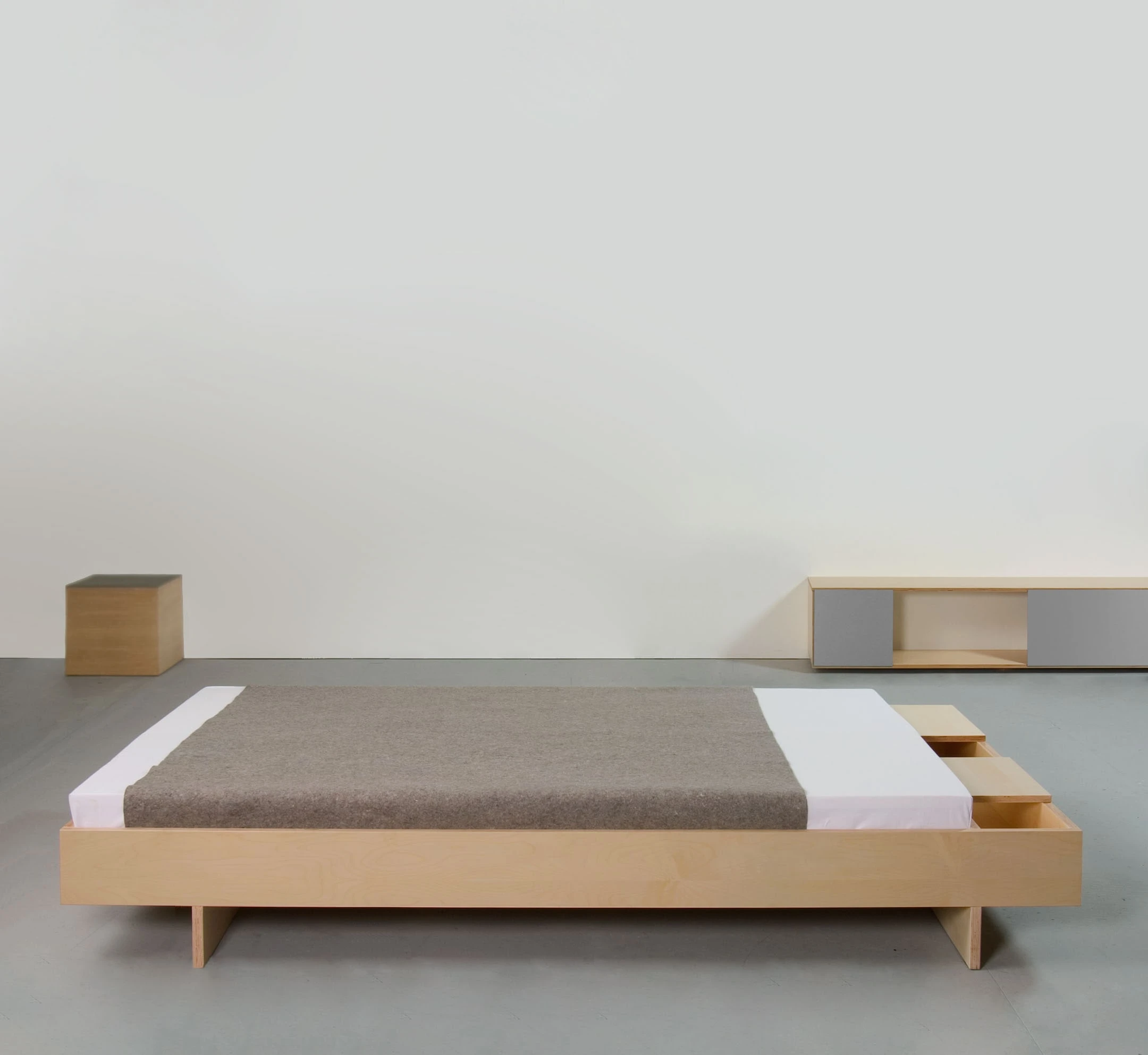 Bett aus Ahorn mit grauer Decke und seitlichem Fach, dahinter ein Lowboard und ein kleiner Holzkubus.