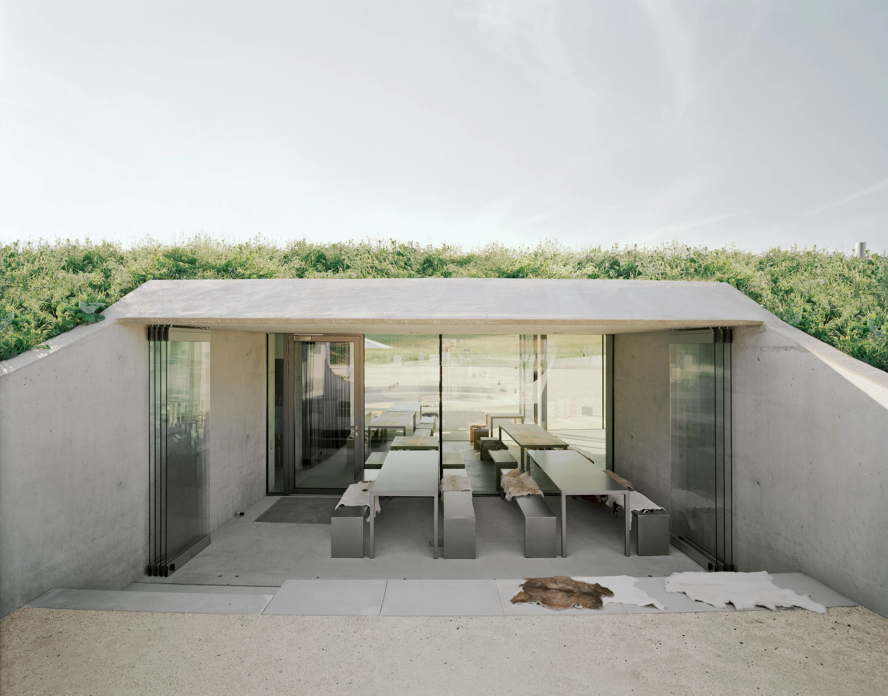 In den Hang gebauter Pavillon mit begrüntem Dach; hinter der offenen Glasfront Tische und Bänke aus Holz.
