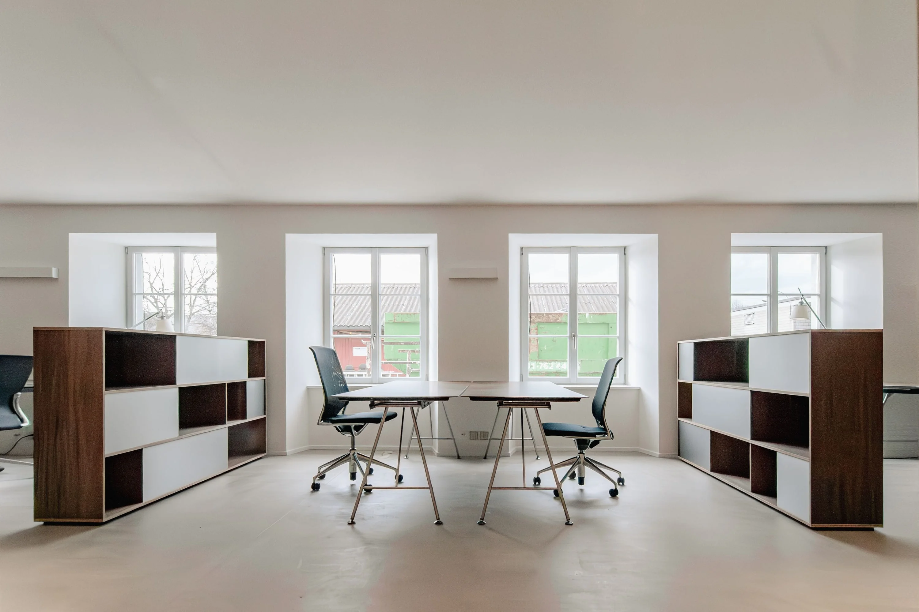 Büroraum mit hohen Fenstern, zwei Highboards aus Nussbaum mit offenen Fächern und Arbeitstischen mit Drehstühlen.
