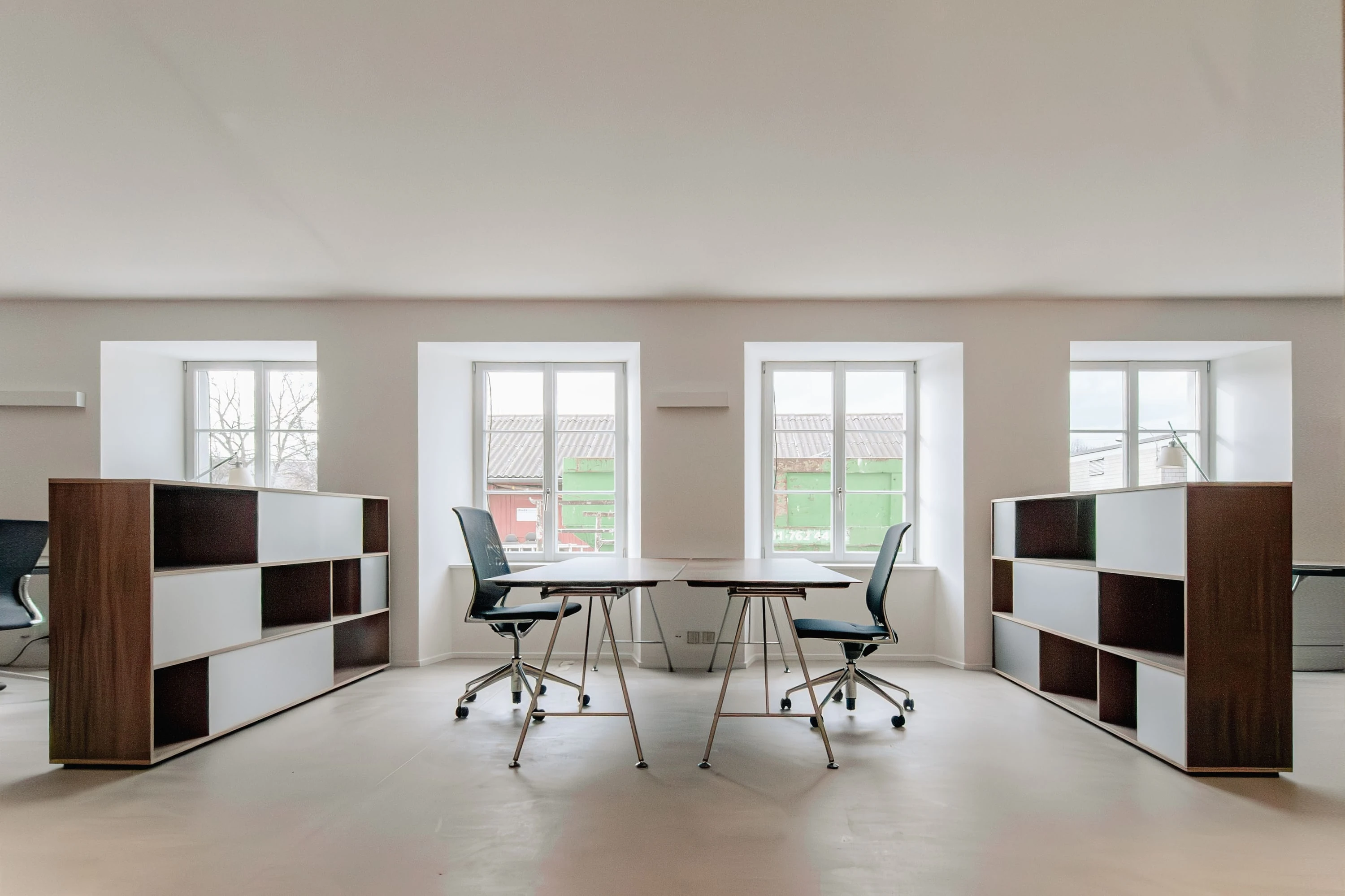 Büroraum mit hohen Fenstern, zwei Highboards aus Nussbaum mit offenen Fächern und Arbeitstischen mit Drehstühlen.