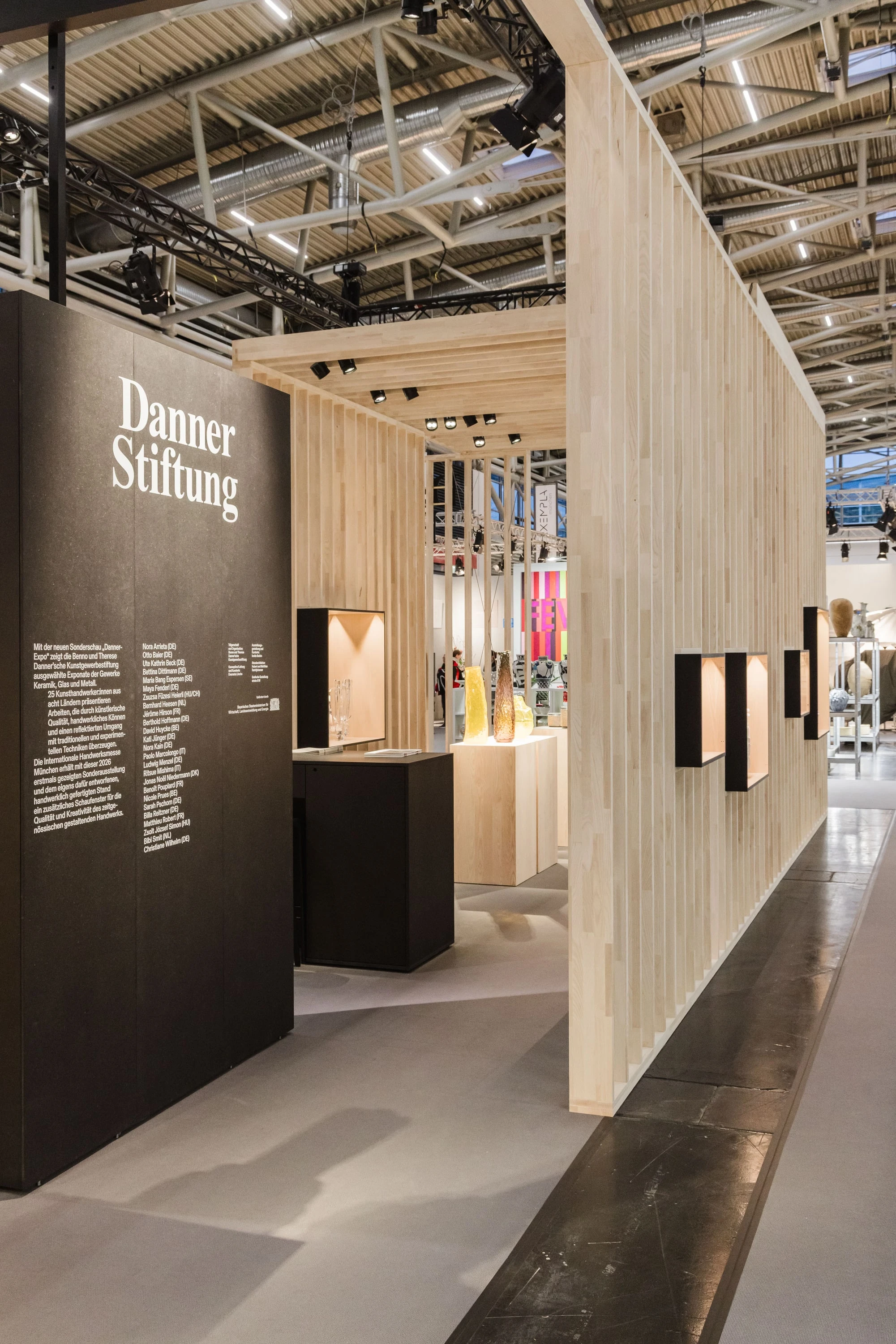 Innenraum des Stands mit schwarzer Texttafel der Danner Stiftung, Holzsockeln mit Objekten und einer Lamellenwand mit Vitrinen.