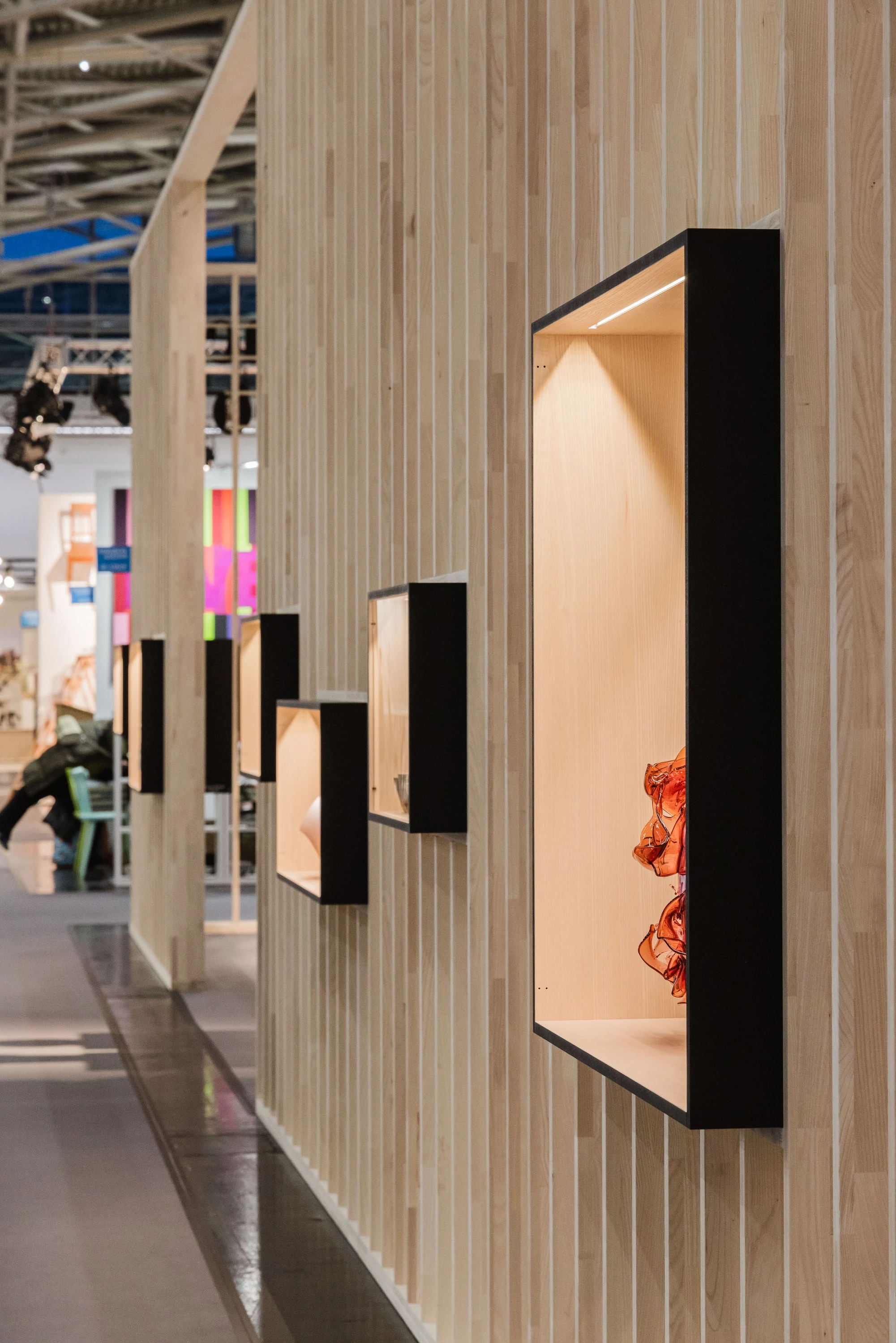 Lamellenwand von aussen mit mehreren schwarz gerahmten Vitrinen, vorne eine Vitrine mit orangem Objekt.