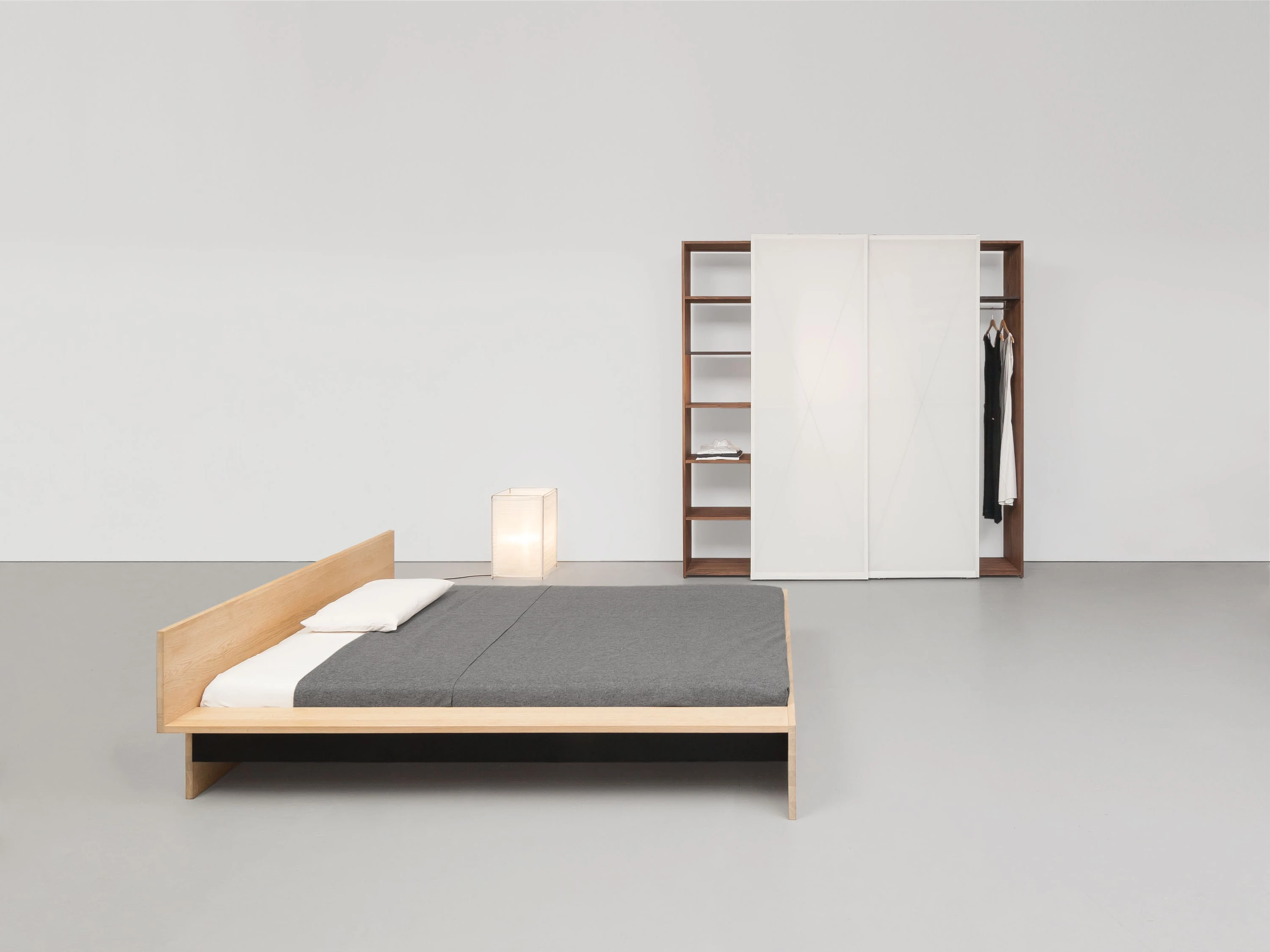 Bett aus Eiche mit Kopfteil und Leuchte, dahinter ein Regal aus Nussbaum und ein weisser Schrank mit Kleidung.