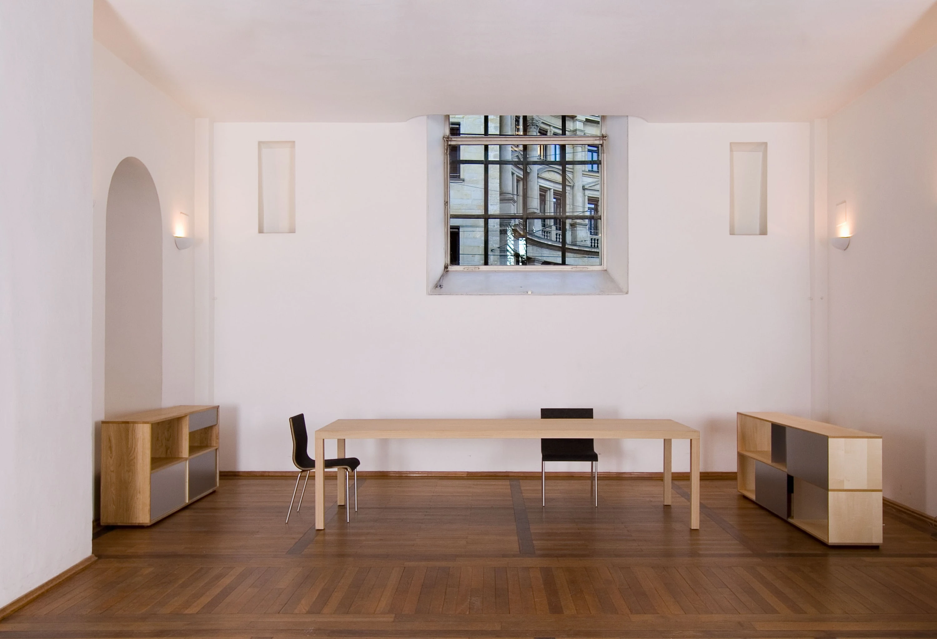 Hoher Raum mit Parkettboden und Rundbogentür, darin ein langer Holztisch mit zwei schwarzen Stühlen und zwei Sideboards.