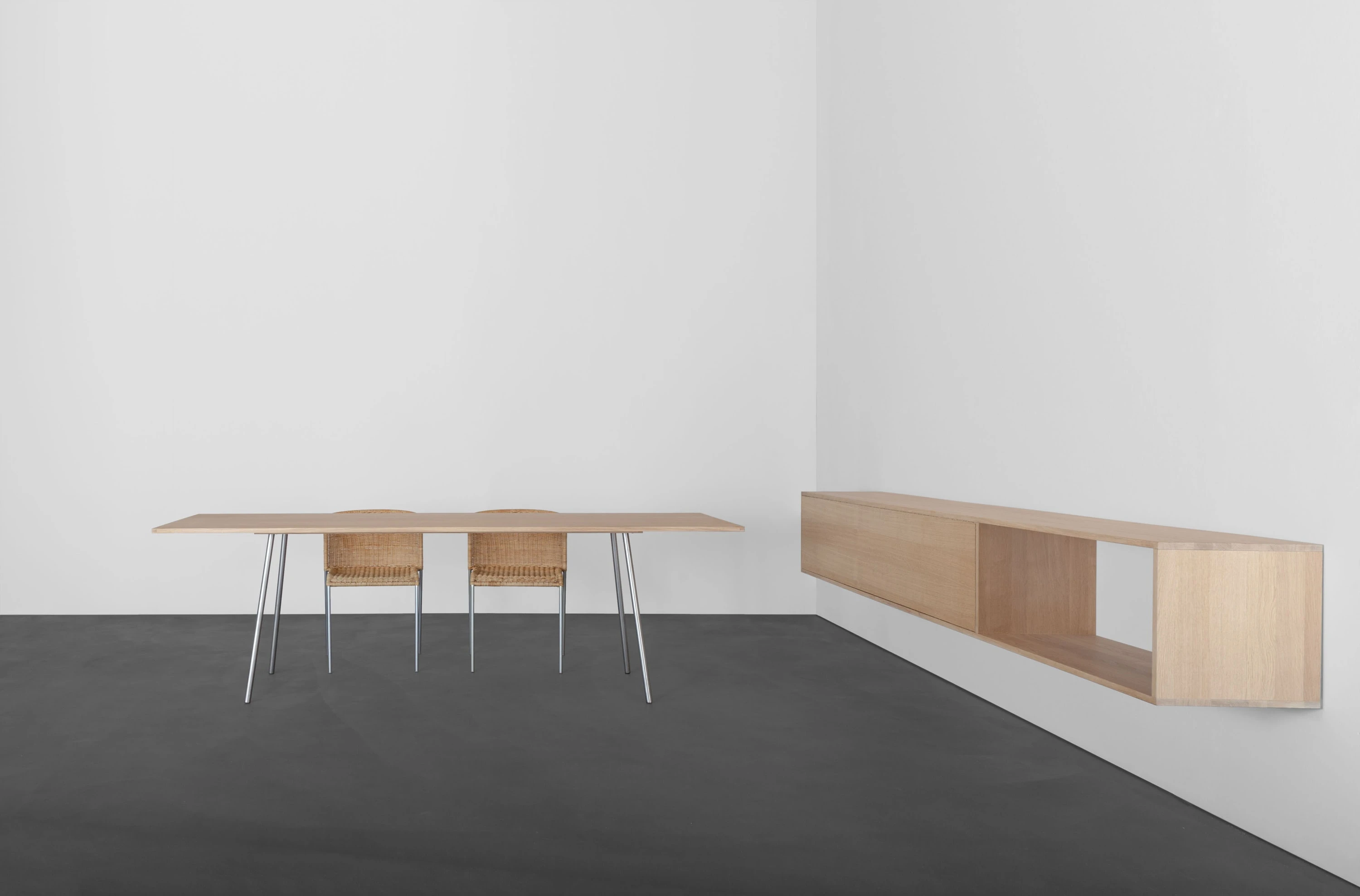 Heller Tisch mit schlanken Metallbeinen und zwei Holzstühlen, rechts ein langes Wandregal aus Holz.