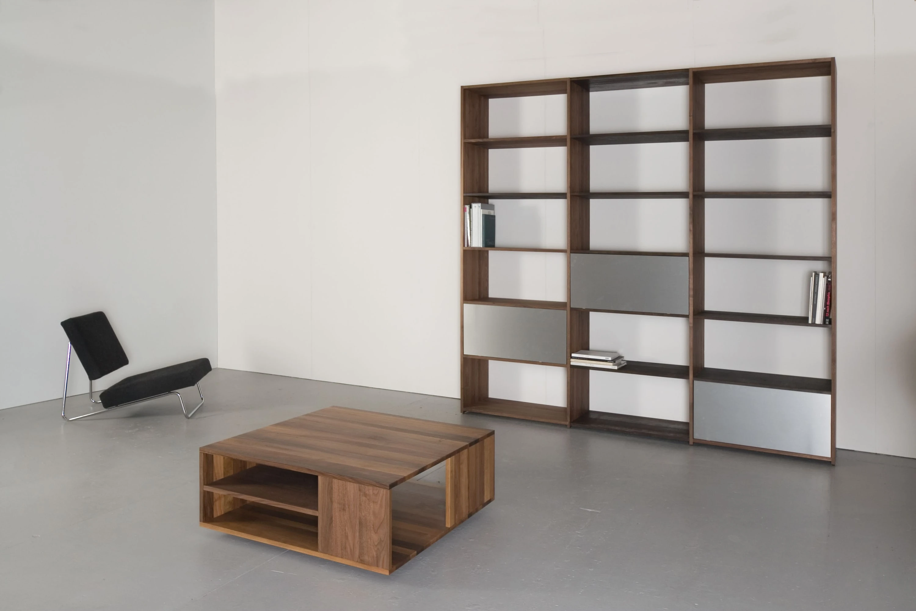 Hohes Regal aus dunklem Holz mit grauen Fronten, davor ein Salontisch aus dunklem Holz und ein schwarzer Liegesessel.