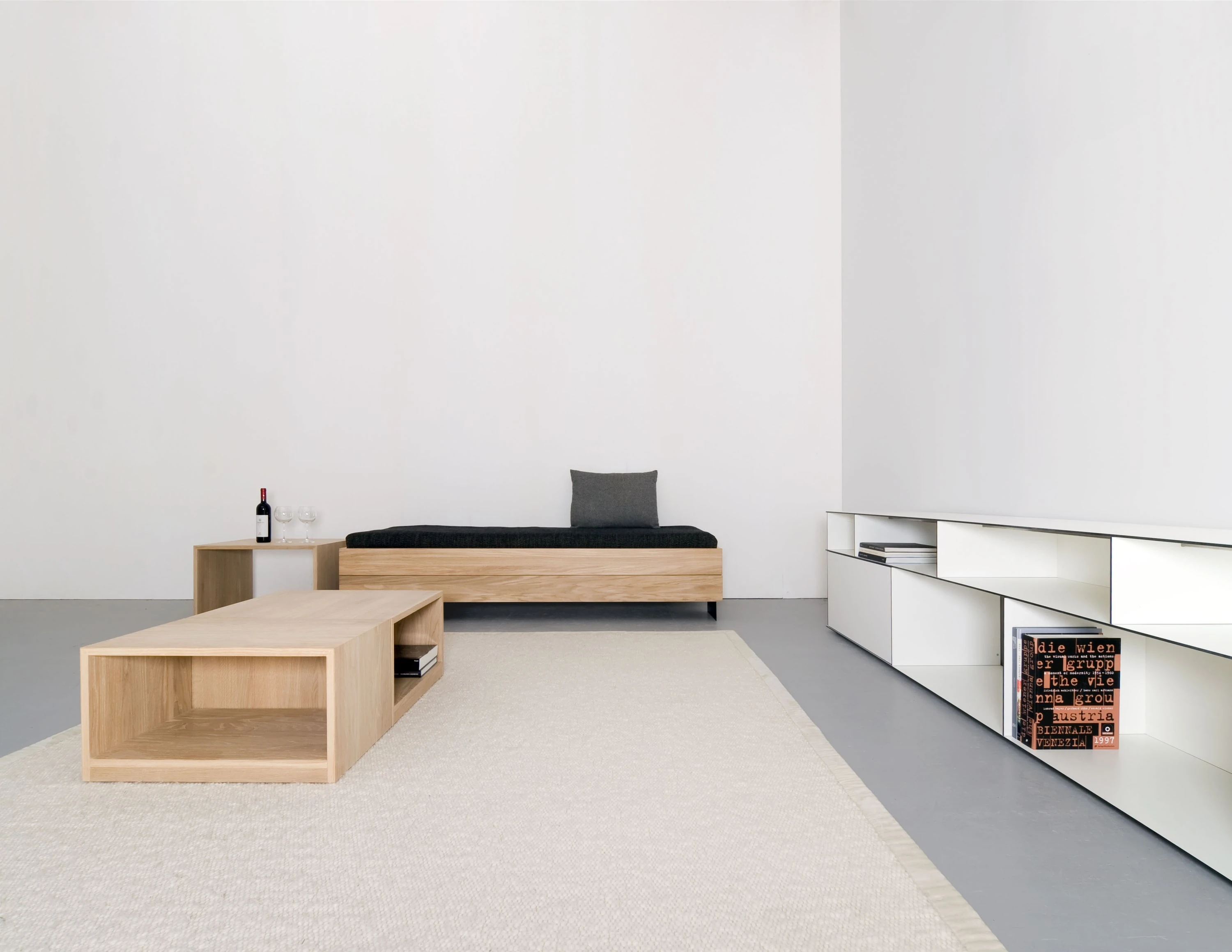 Niedriges Bett mit schwarzer Decke, davor ein offener Holzkasten als Salontisch auf hellem Teppich, rechts ein langes weisses Regal mit Büchern.