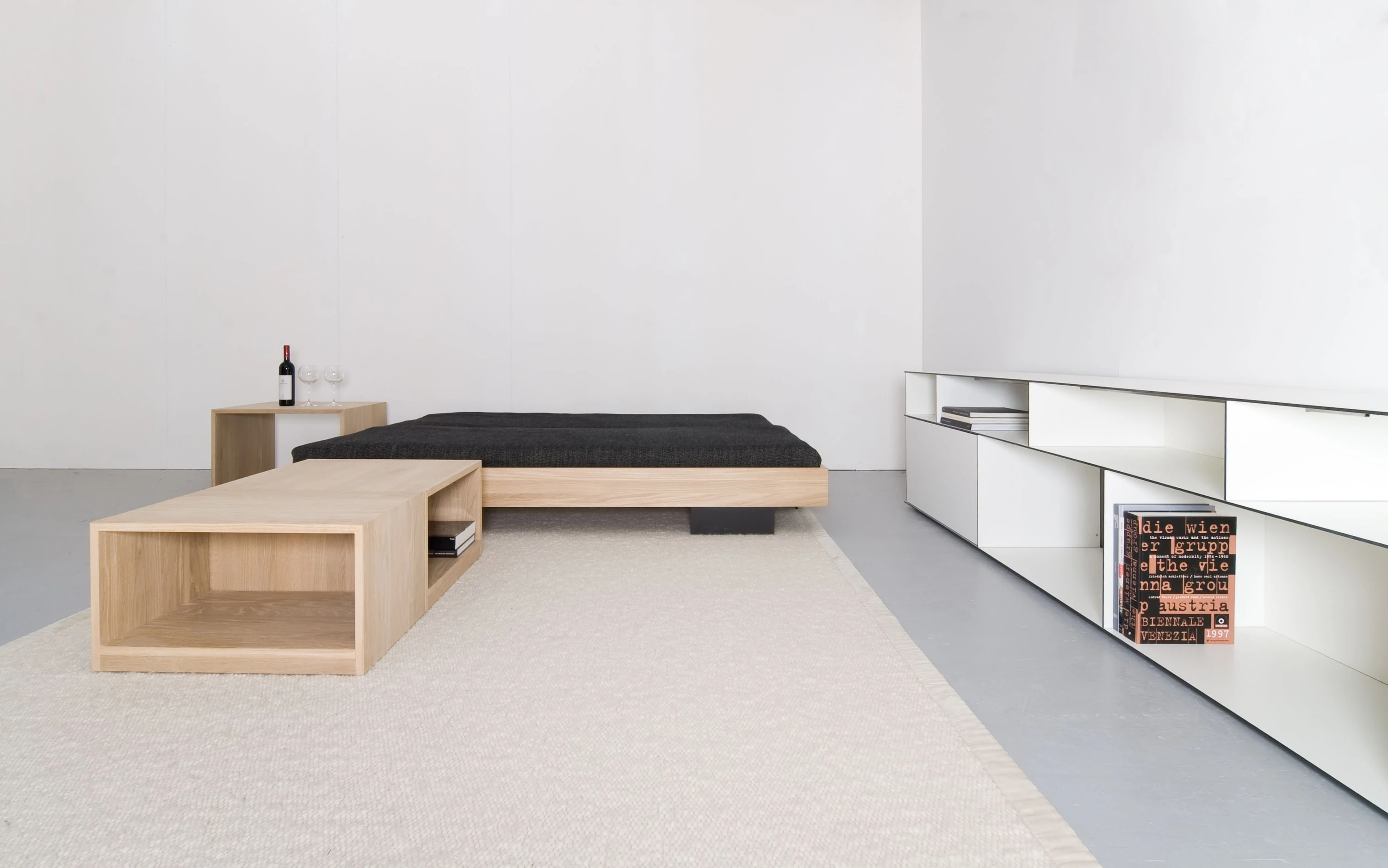 Bett mit schwarzer Decke hinter einem offenen Holzkasten auf hellem Teppich, rechts ein langes weisses Regal mit Büchern.