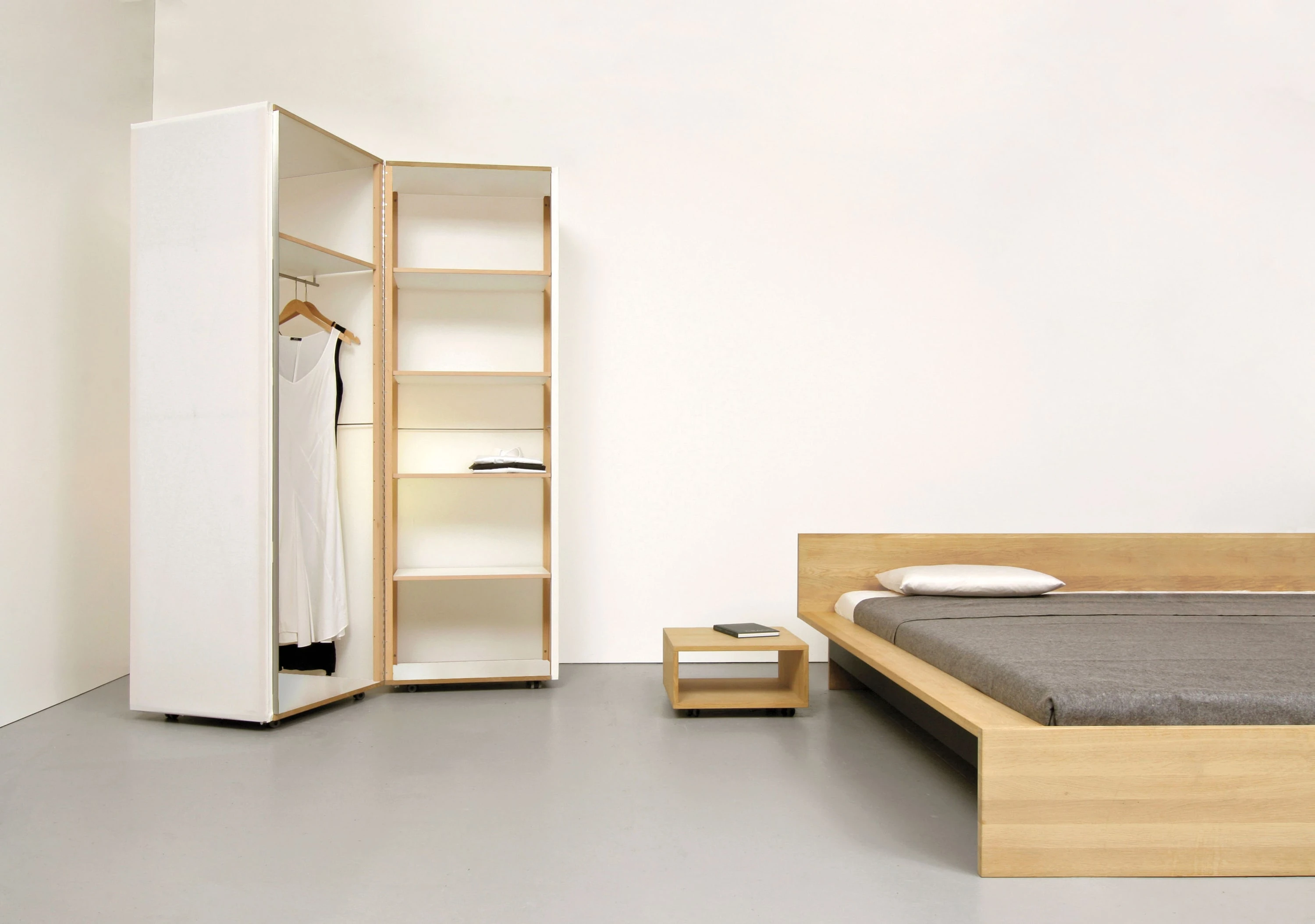 Geöffneter weisser Textilschrank mit Kleid, daneben ein Bett aus Eiche und ein kleiner Holzkubus.