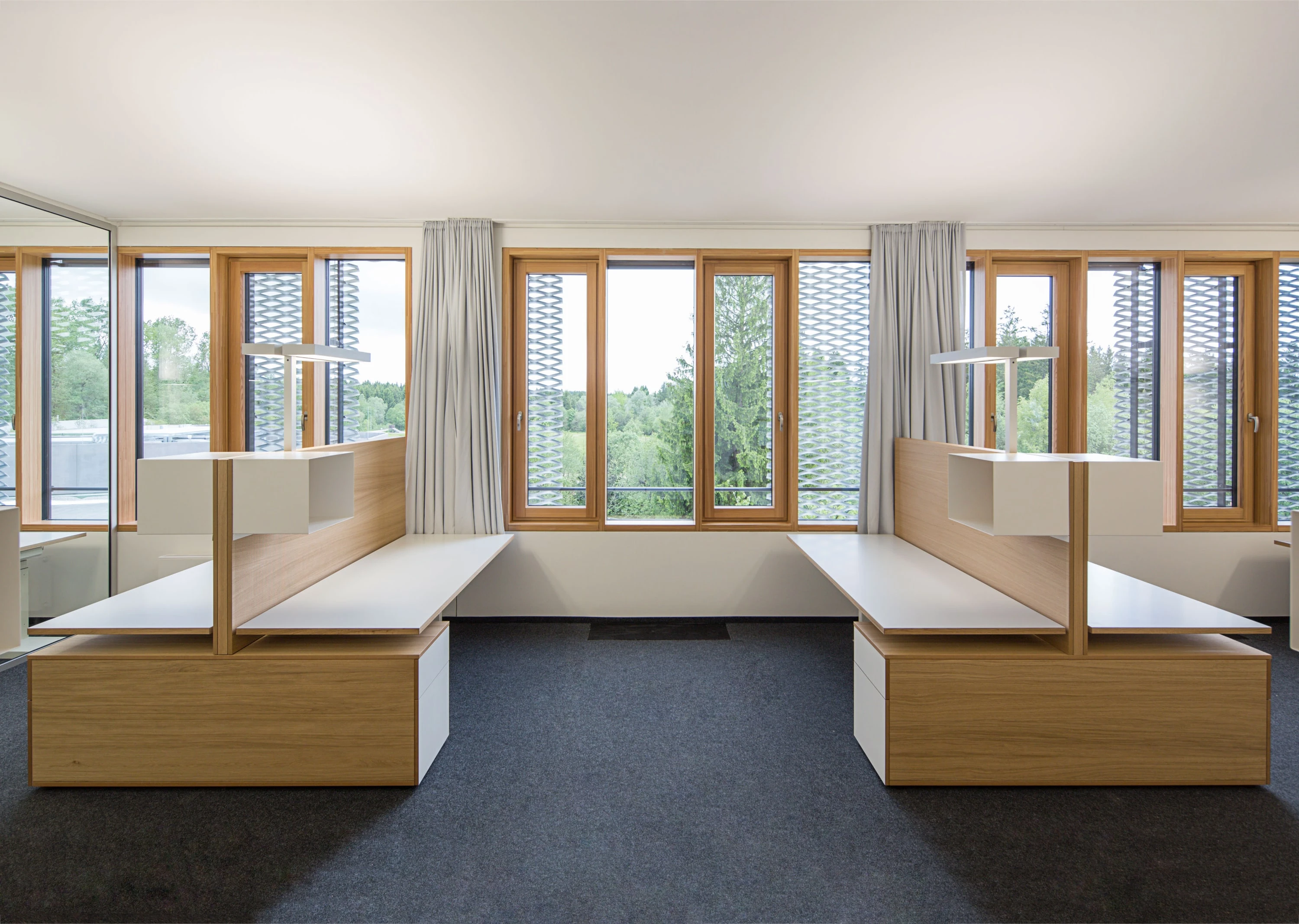 Workstations with tall wooden partitions and white desktops in front of a row of windows.