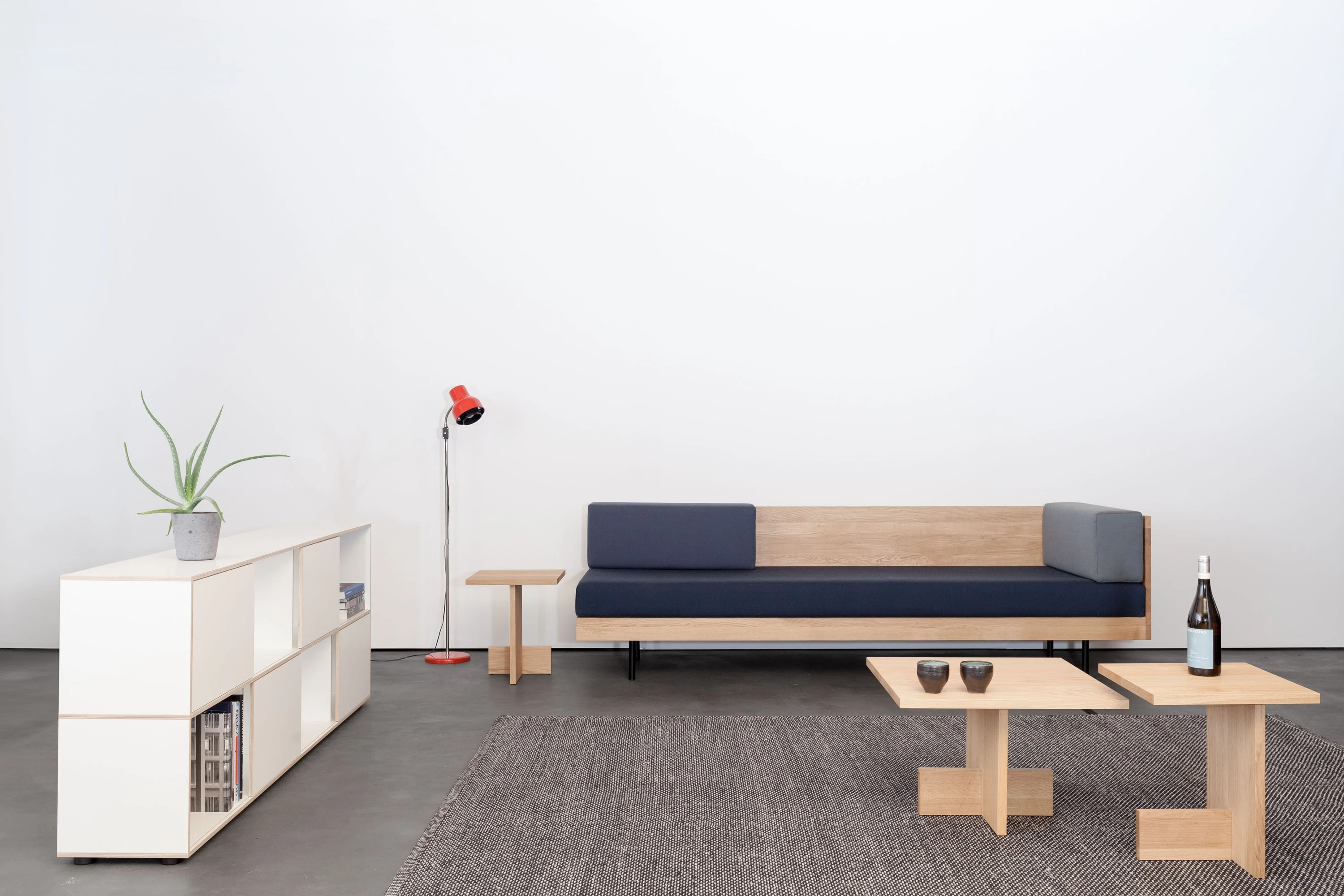 Couch aus Eiche mit blauen Polstern, davor niedrige Beistelltische aus Eiche auf grauem Teppich, links ein weisses Sideboard mit Pflanze und eine rote Leuchte.