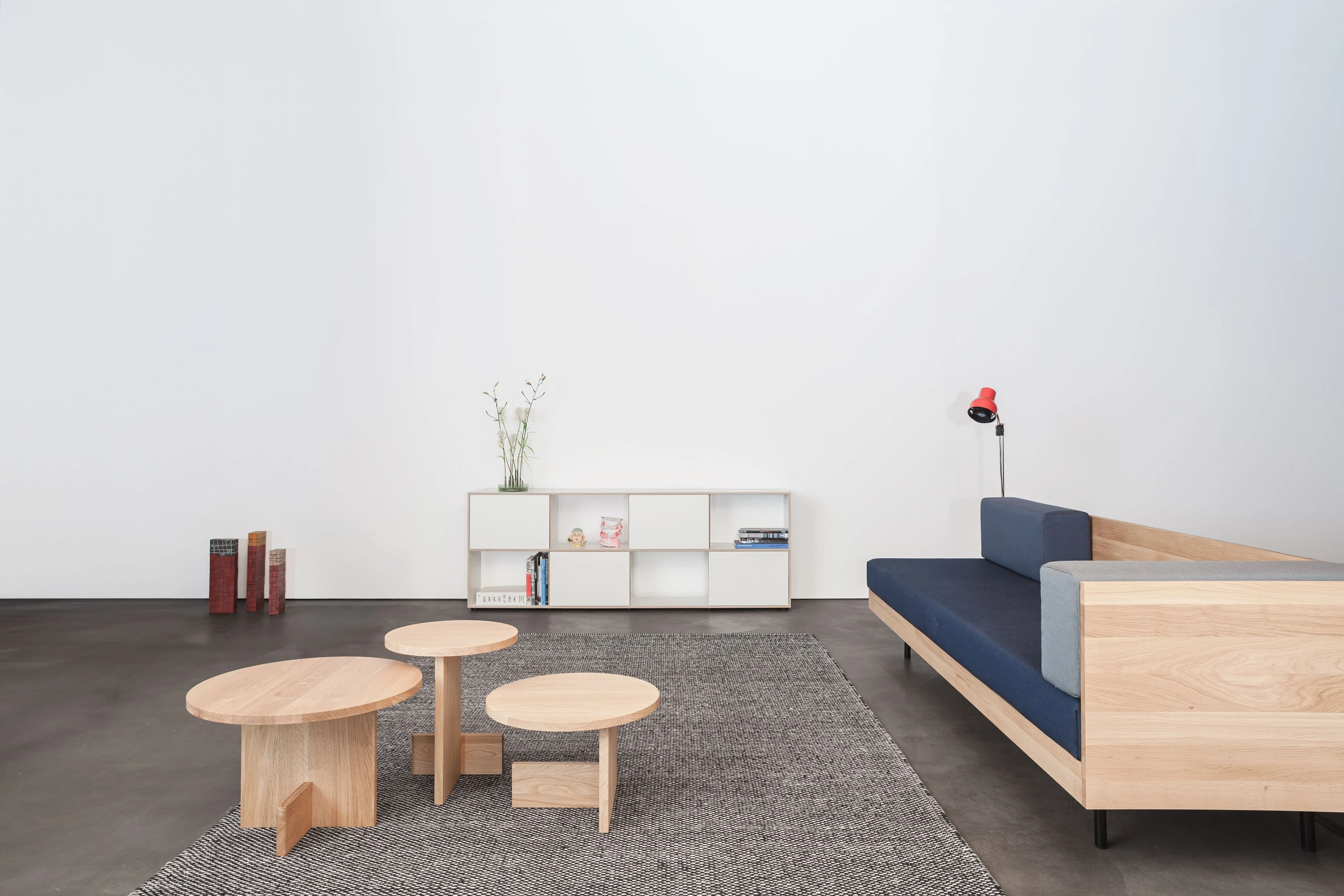 Drei runde Beistelltische aus Eiche auf grauem Teppich, dahinter ein weisses Sideboard, rechts eine Couch mit blauen Polstern.