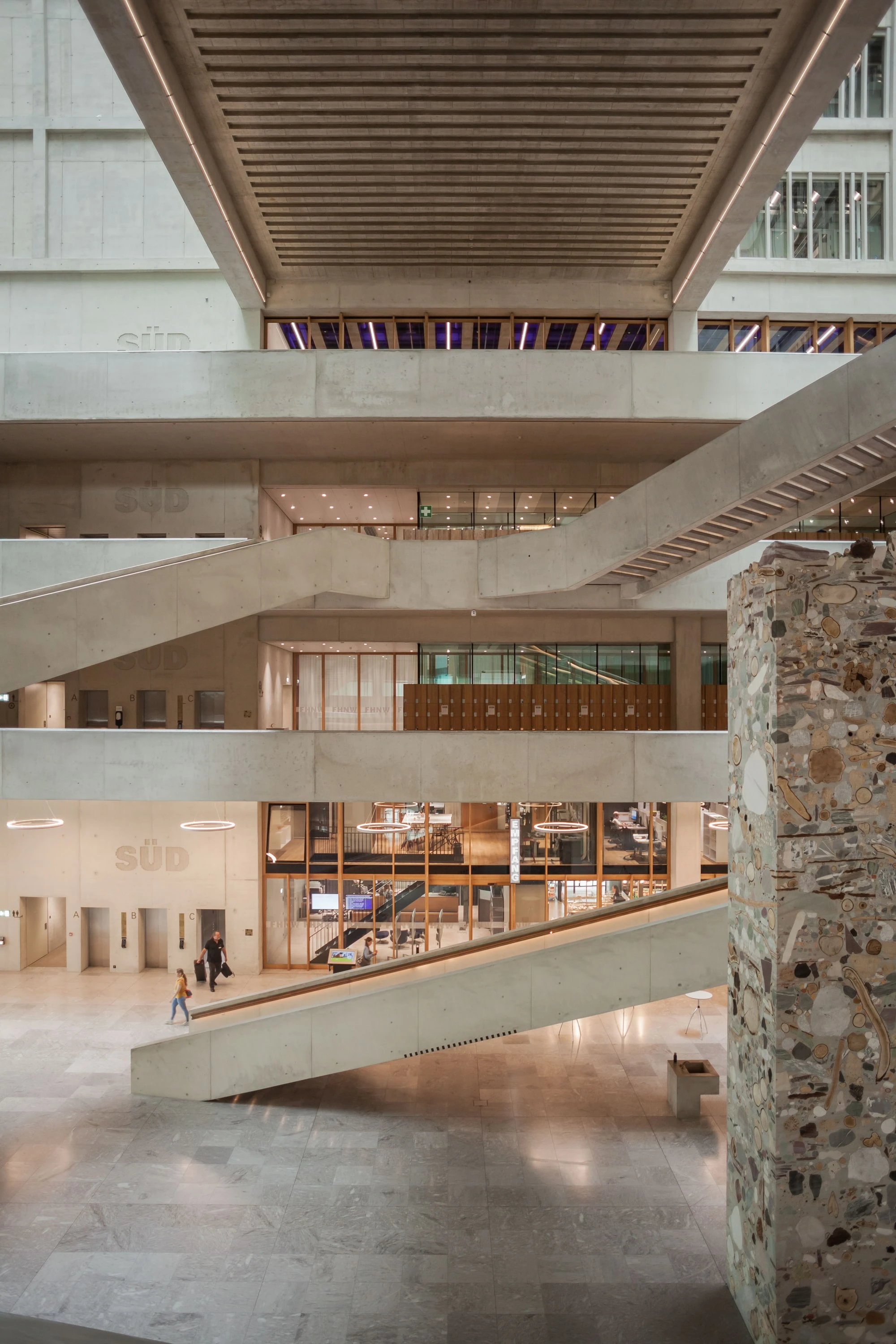 Blick durch das mehrgeschossige Atrium mit diagonalen Treppen und einer Steinwand.