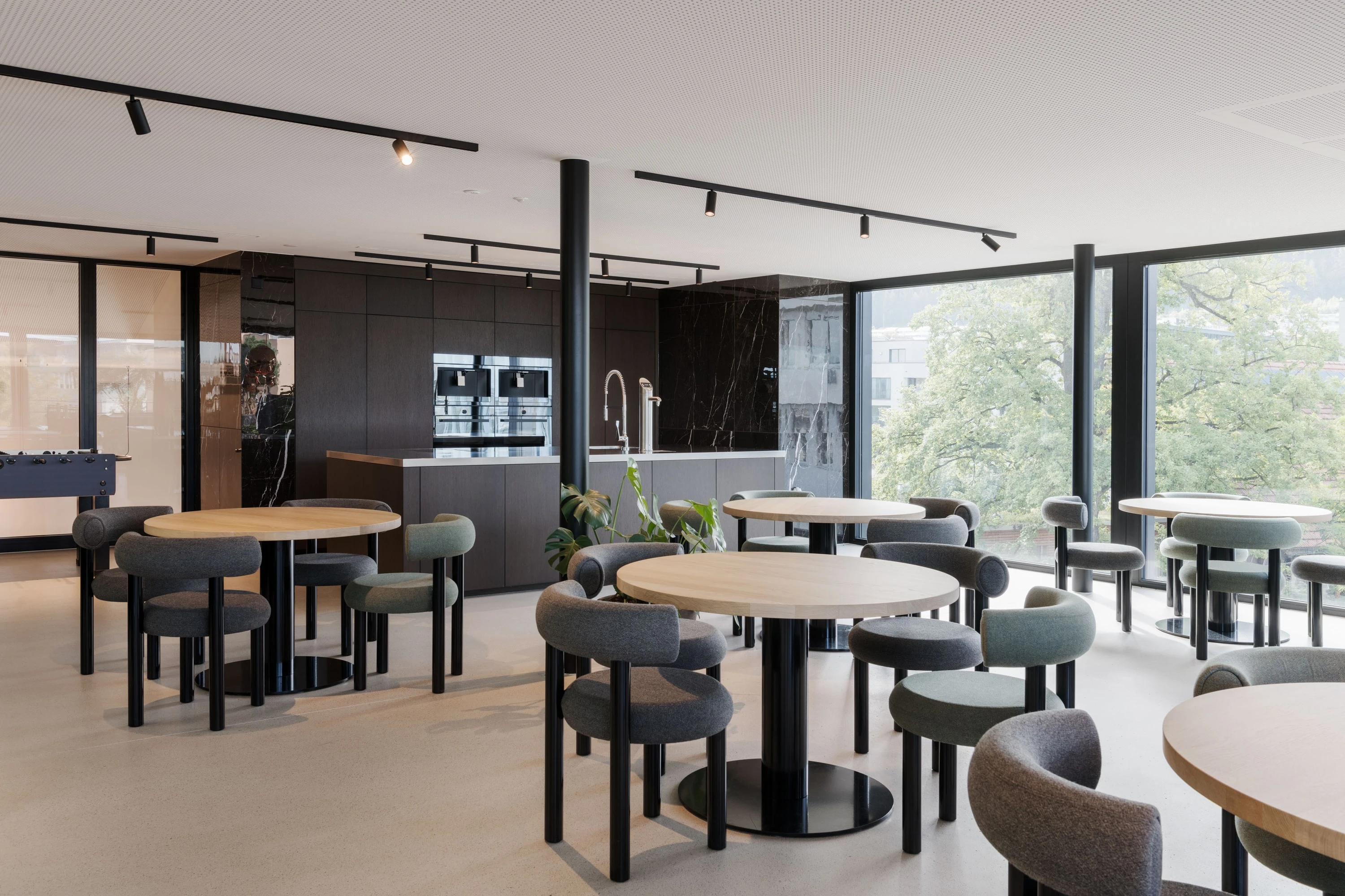Cafeteria mit runden Tischen, grauen Sesseln und Küchenzeile vor grosser Verglasung.