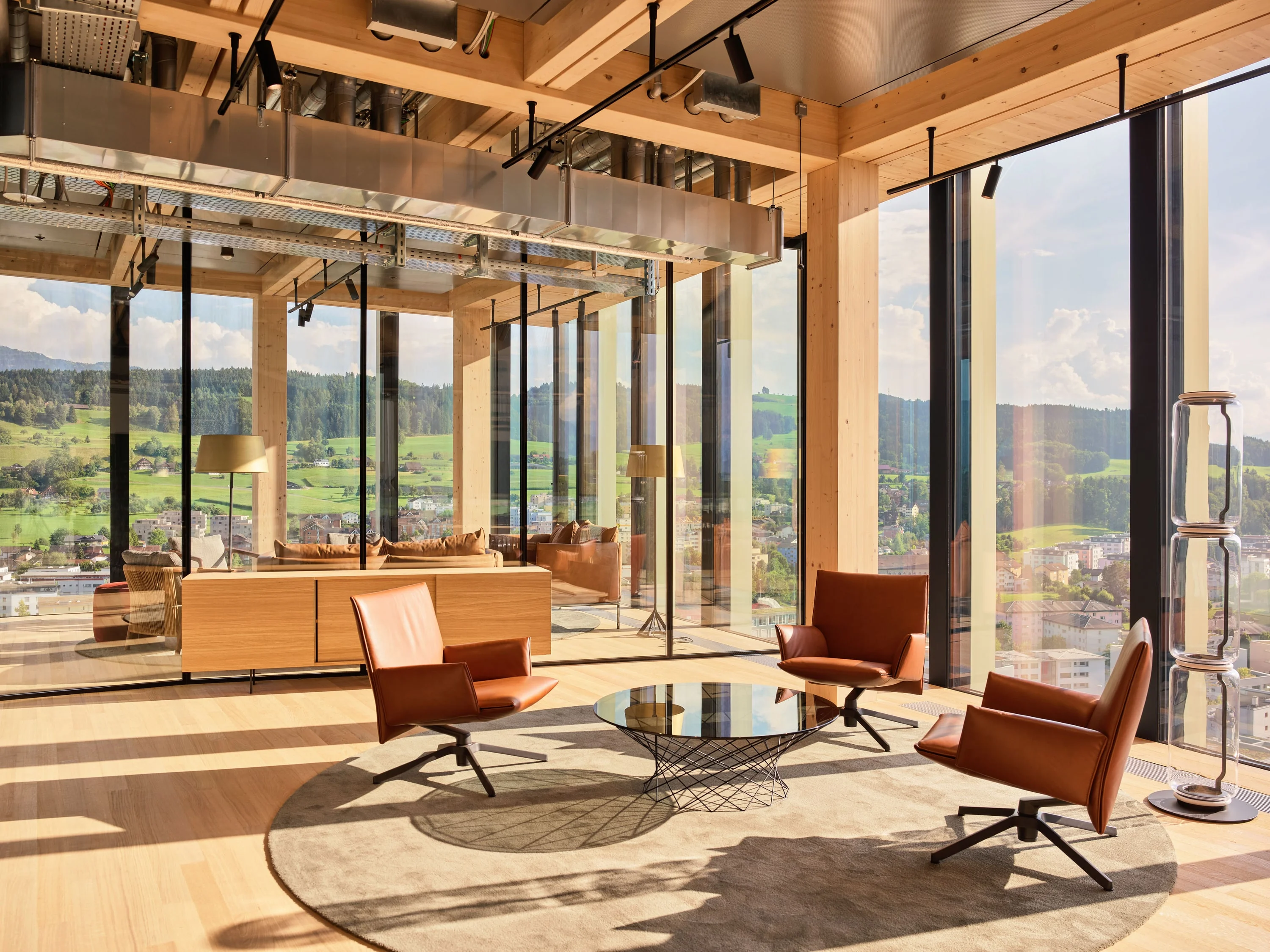 Lounge im Holzbau mit orangen Sesseln, rundem Glastisch und Blick über die Landschaft.