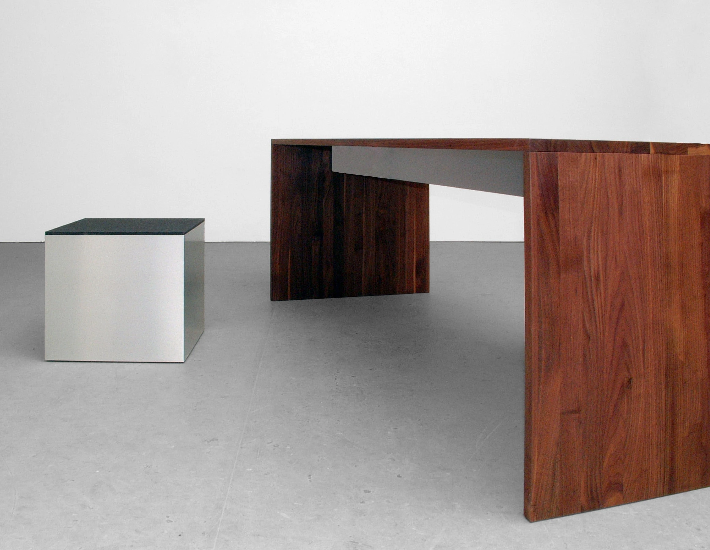 Tisch mit breiten Seitenwangen aus dunklem Holz und Metallzarge, daneben ein Metallkubus.