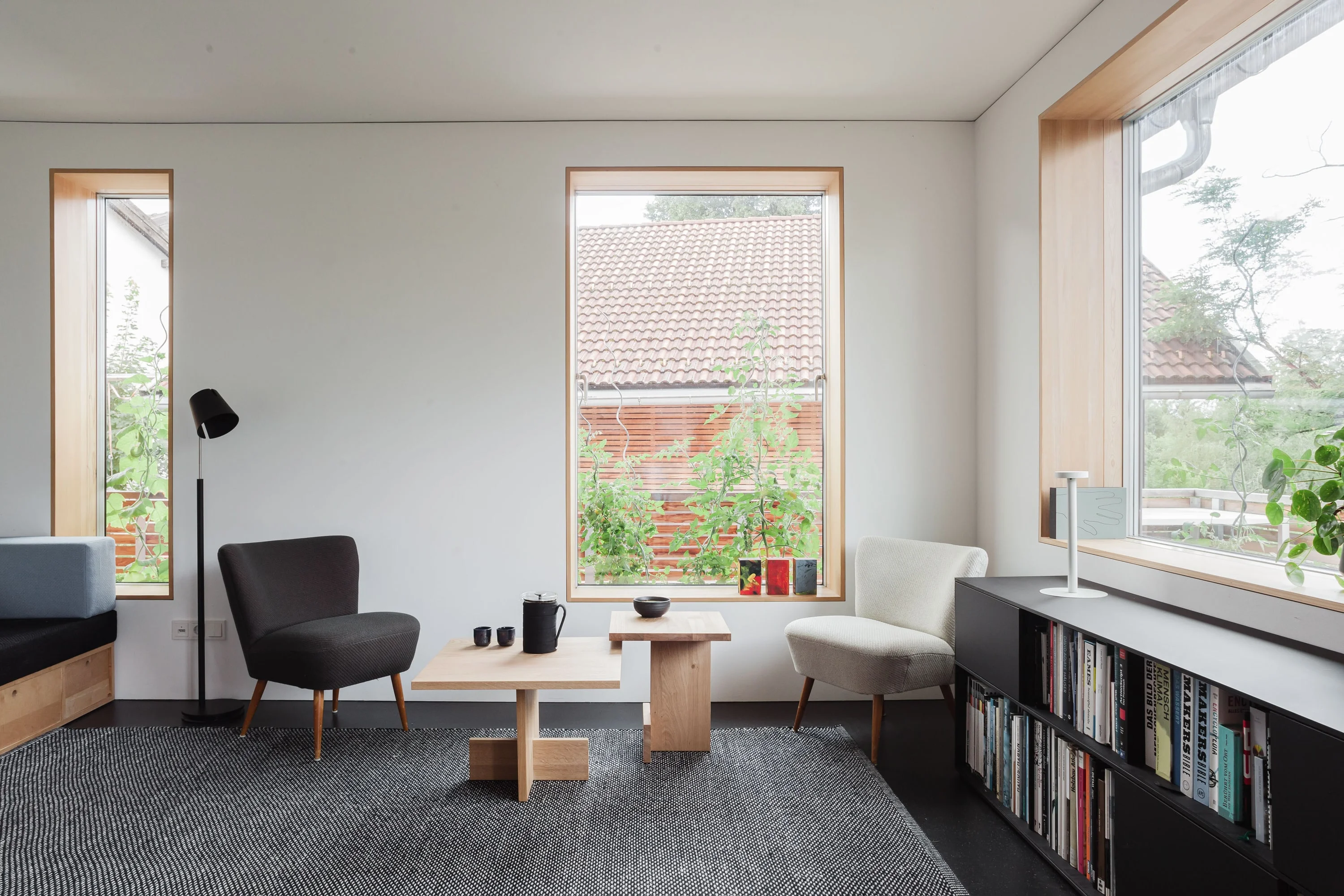Wohnzimmer mit Sesseln, niedrigem Holztisch, Bücherregal unter dem Fenster und grossen Fensteröffnungen.
