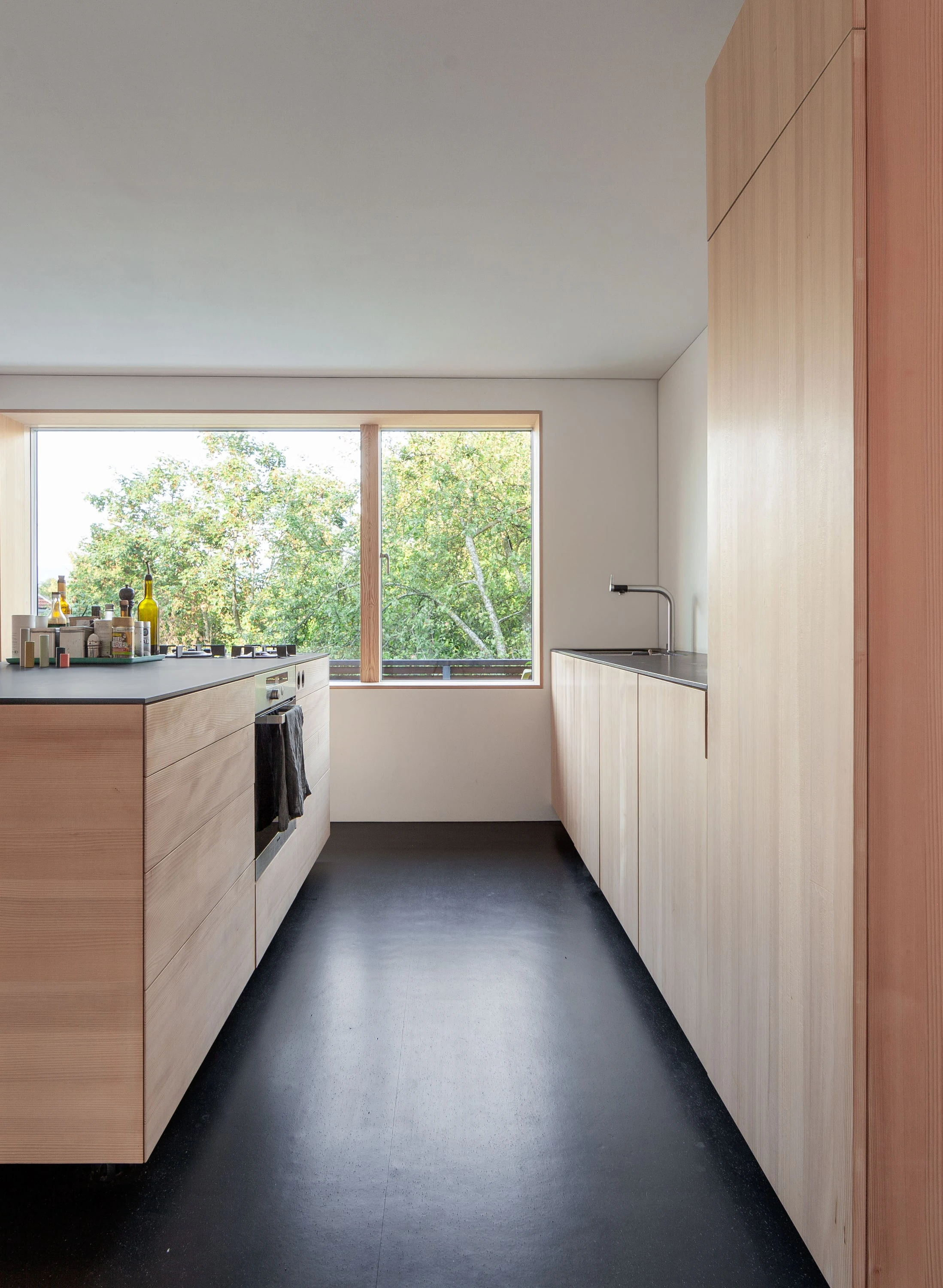 Schmale Küche mit hellen Holzfronten, dunklem Boden und Fenster am Ende.