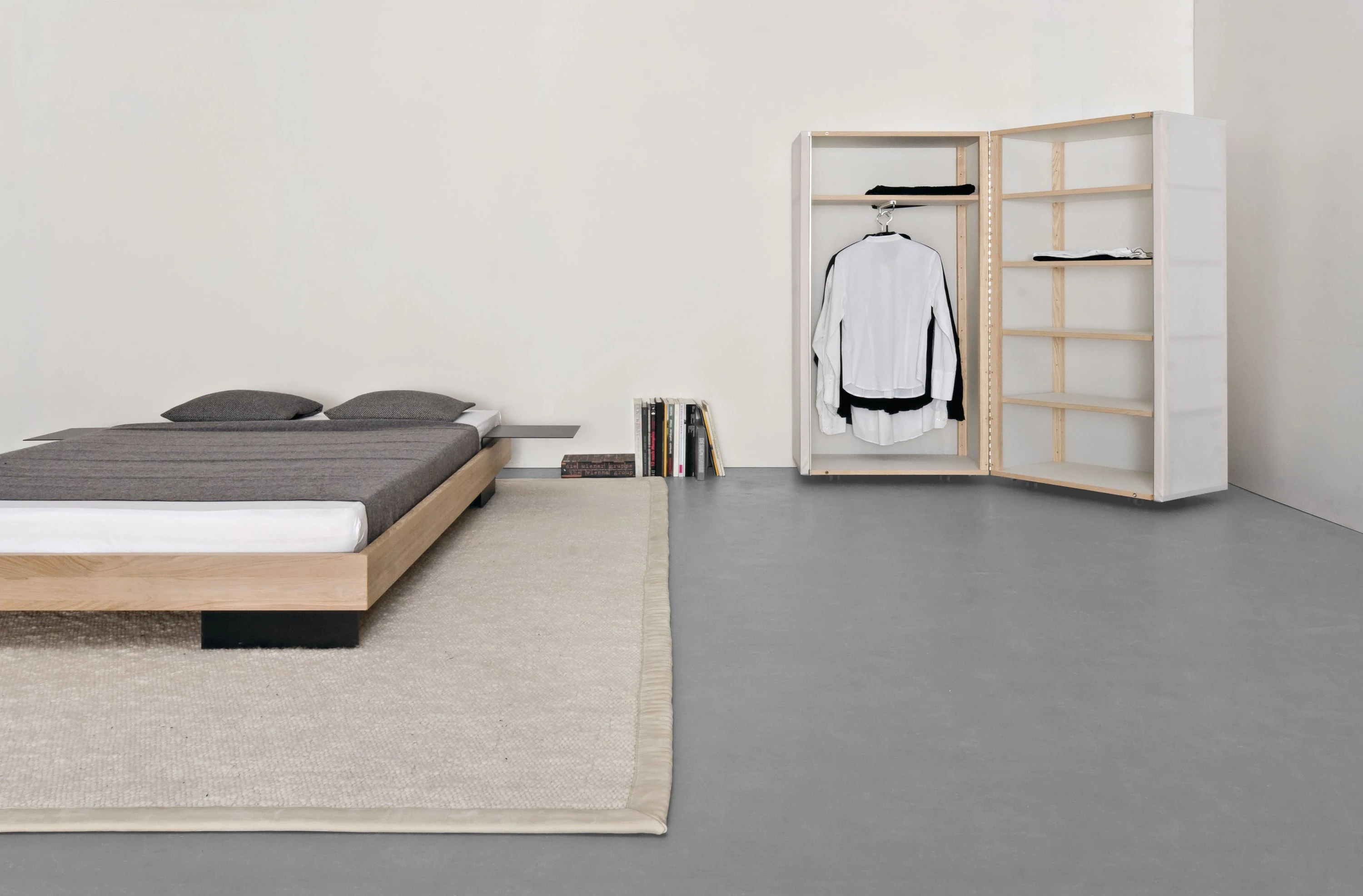 Bett aus Eiche auf hellem Teppich, dahinter ein geöffneter Rollschrank mit Hemd und gestapelten Büchern.