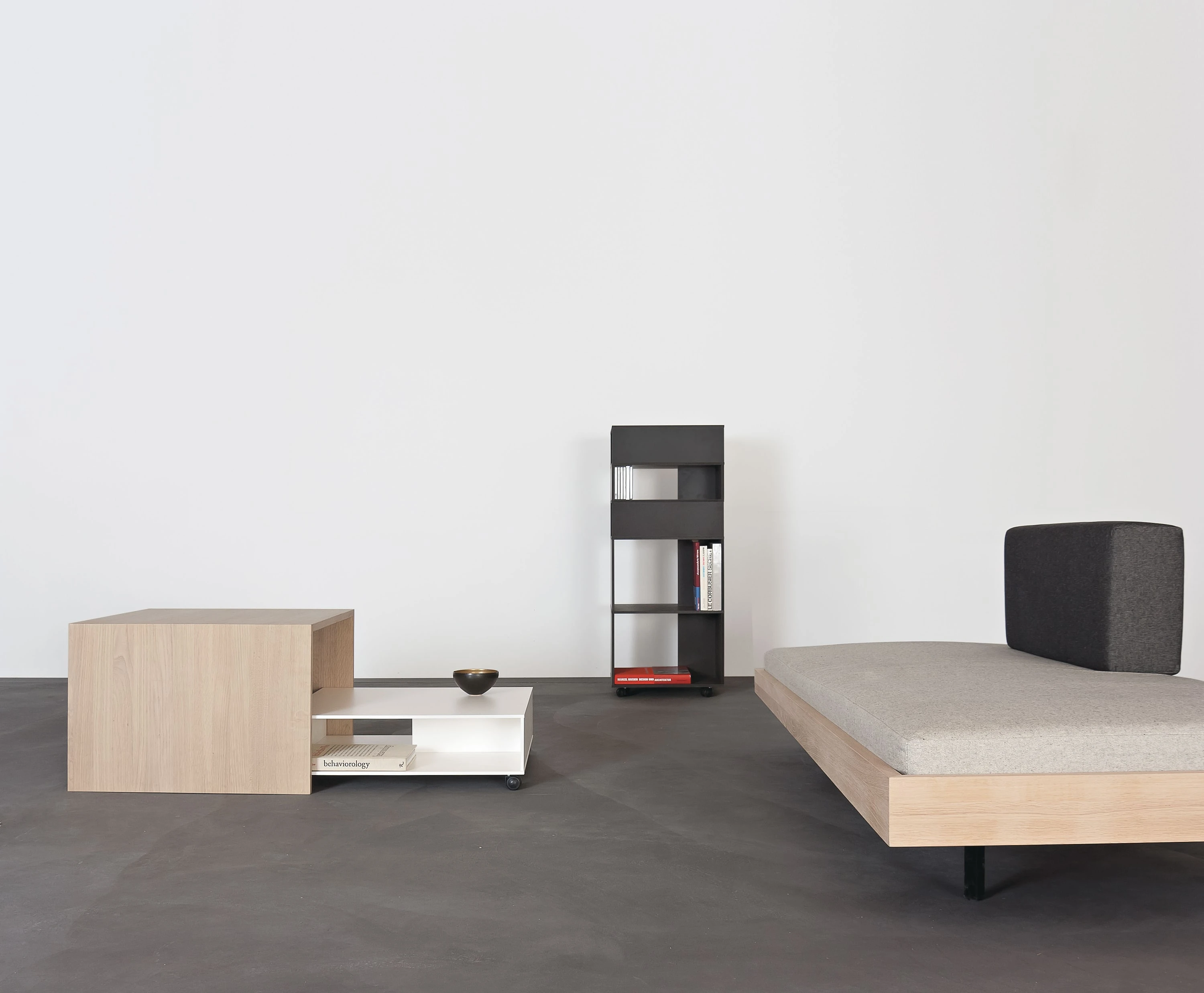 Salontisch aus Eiche und weisser Salontisch, dahinter ein schmales schwarzes Bücherregal und ein Bett mit dunklem Kissen.