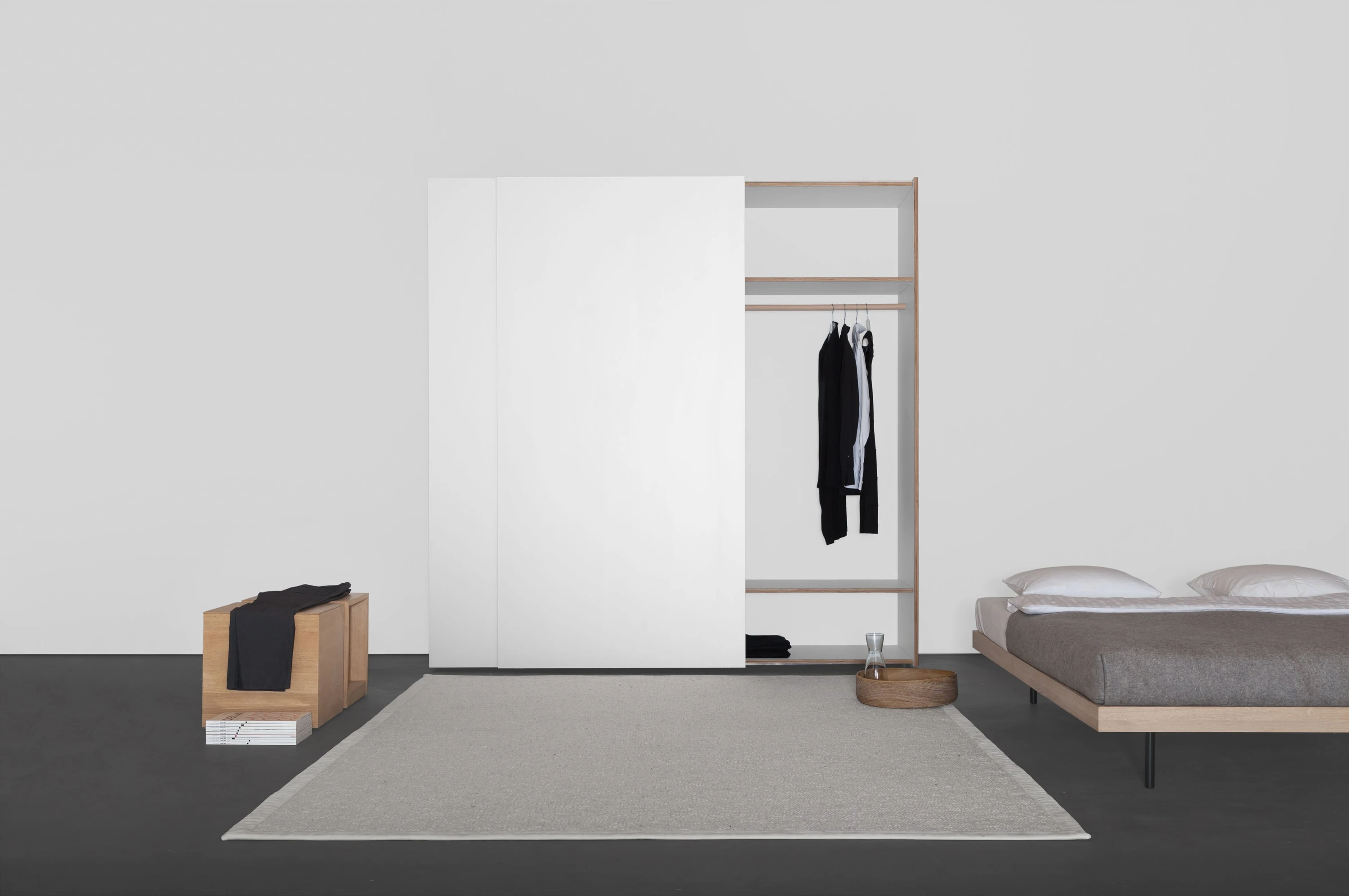 Weisser Kleiderschrank mit geöffneter Schiebetür, davor ein Holzhocker, rechts ein Bett auf hellem Teppich.