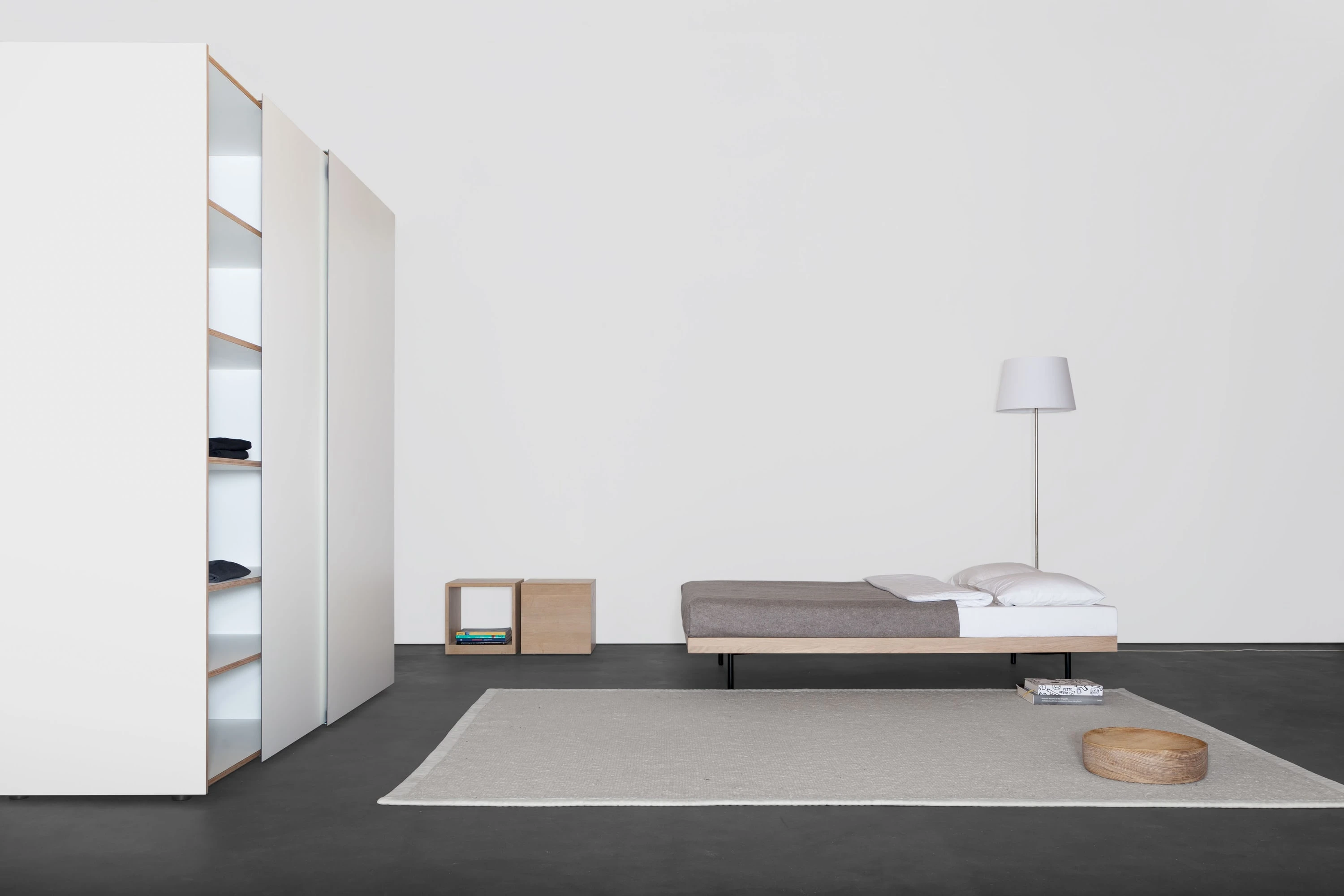 Hoher weisser Kleiderschrank mit offenen Fächern, ein Holzhocker und ein flaches Bett mit Leuchte auf hellem Teppich.