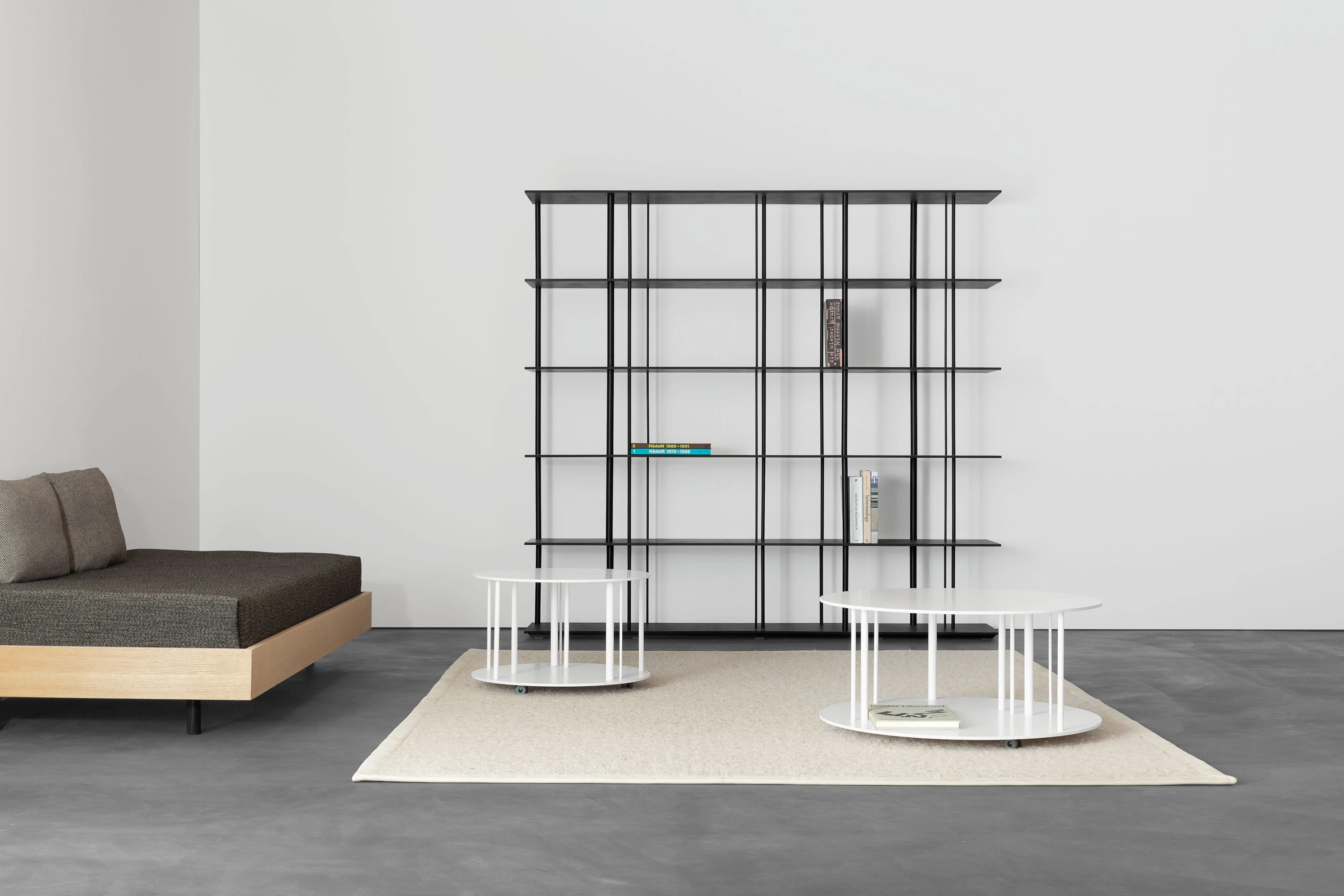 Tagesbett mit dunkler Auflage, schwarzes Metallregal und zwei weisse runde Salontische auf hellem Teppich.
