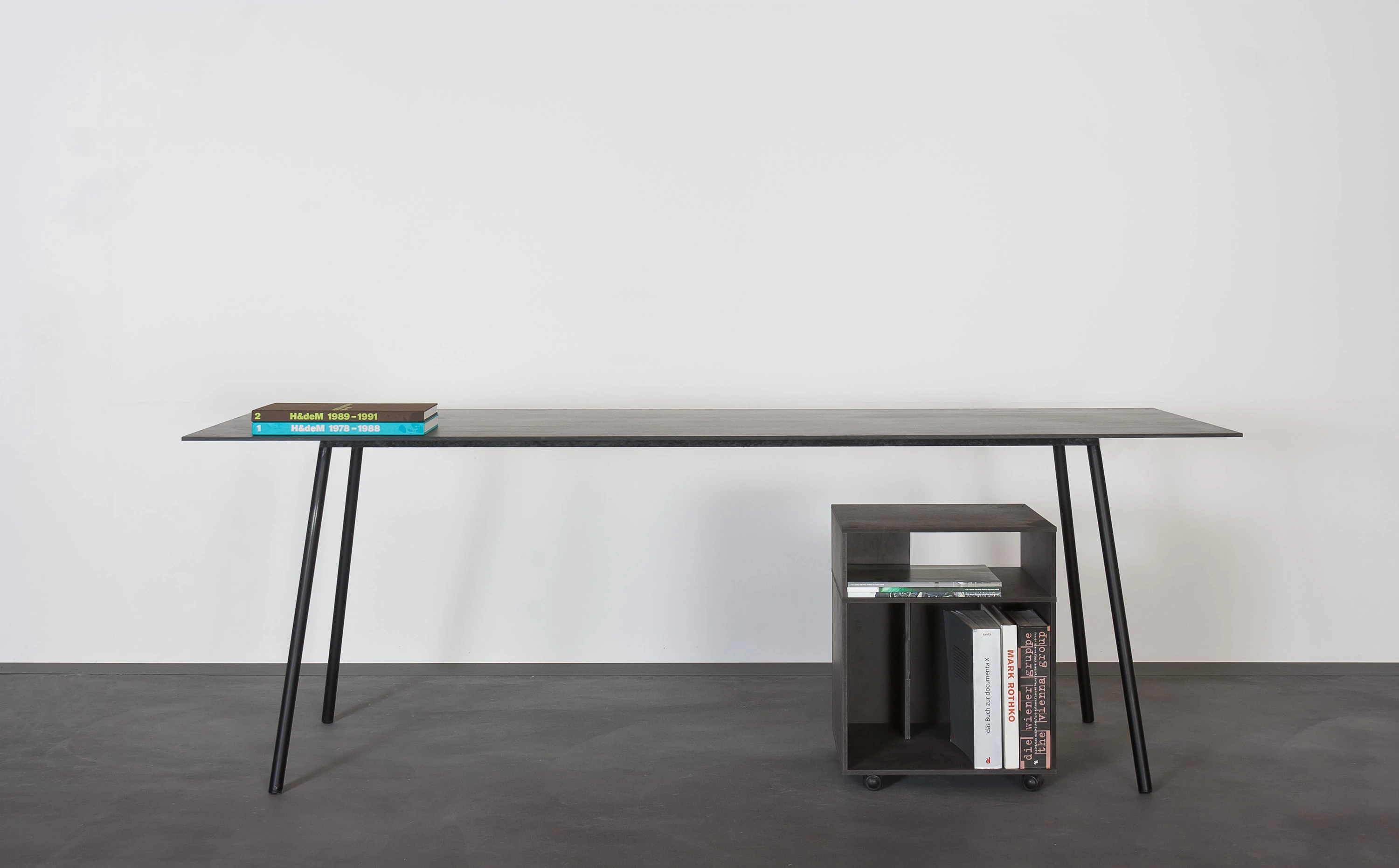 Schwarzer Tisch mit schlanken Beinen und ein schwarzer Rollcontainer mit Büchern.