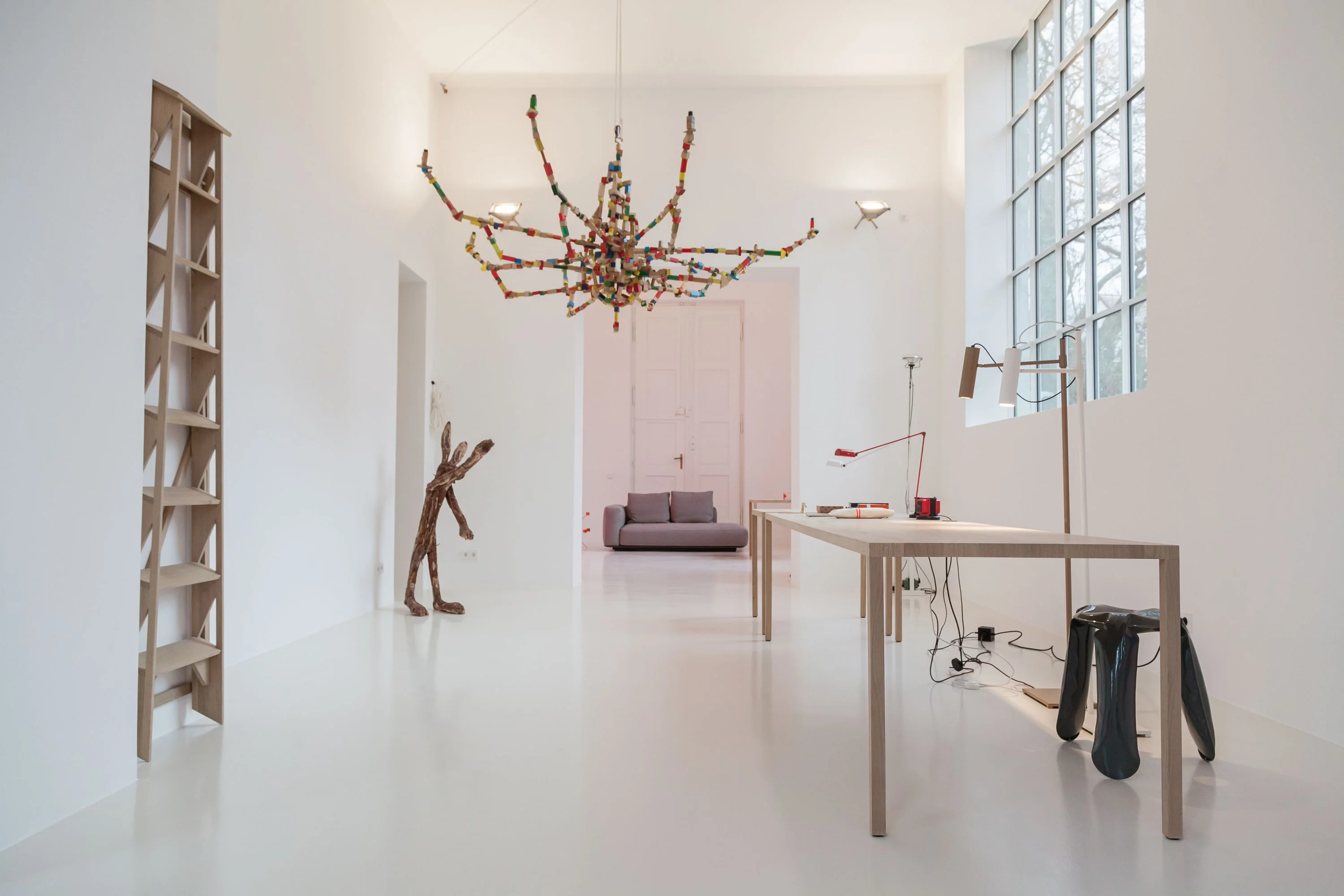 Weisser Ausstellungsraum mit Holztisch, Skulpturen, Leiterregal und Lichtobjekt an der Decke.