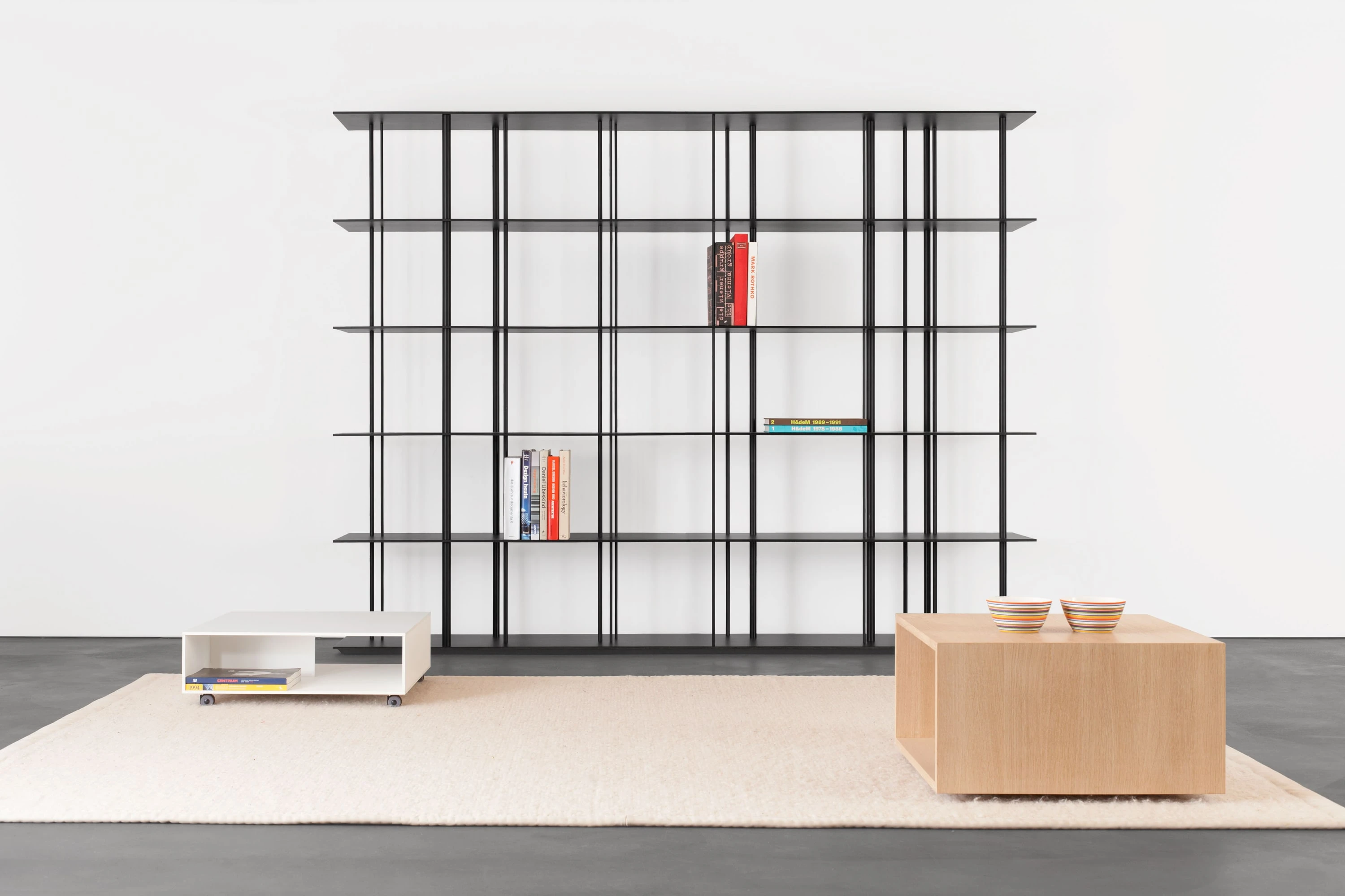 Schwarzes Metallregal mit Büchern, davor ein weisser Salontisch und ein Holzkubus mit Schalen auf hellem Teppich.