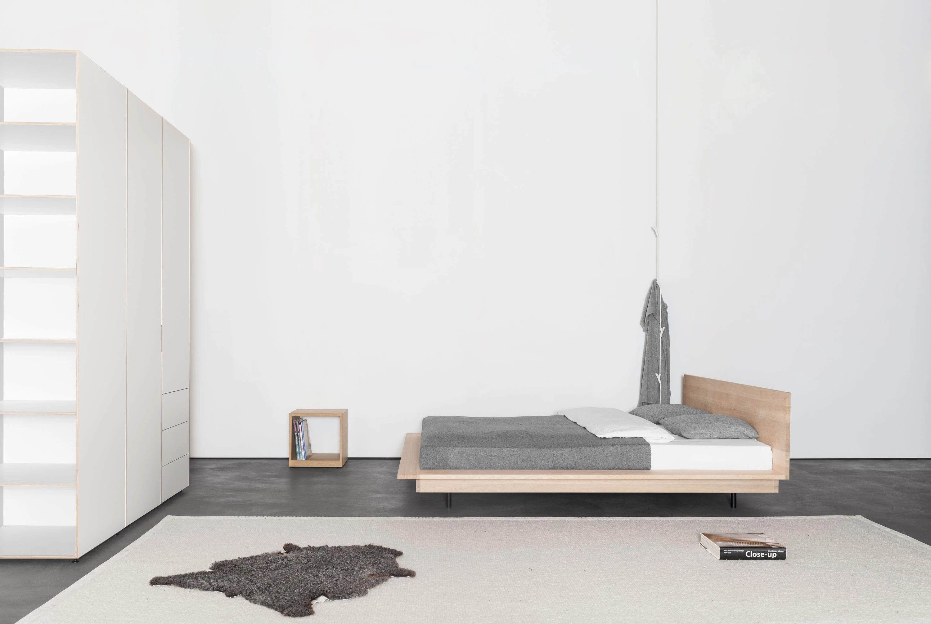 Schlafzimmer mit weissem Kleiderschrank, Bett aus Eiche, kleinem Holzkubus und aufgehängtem Kleidungsstück.