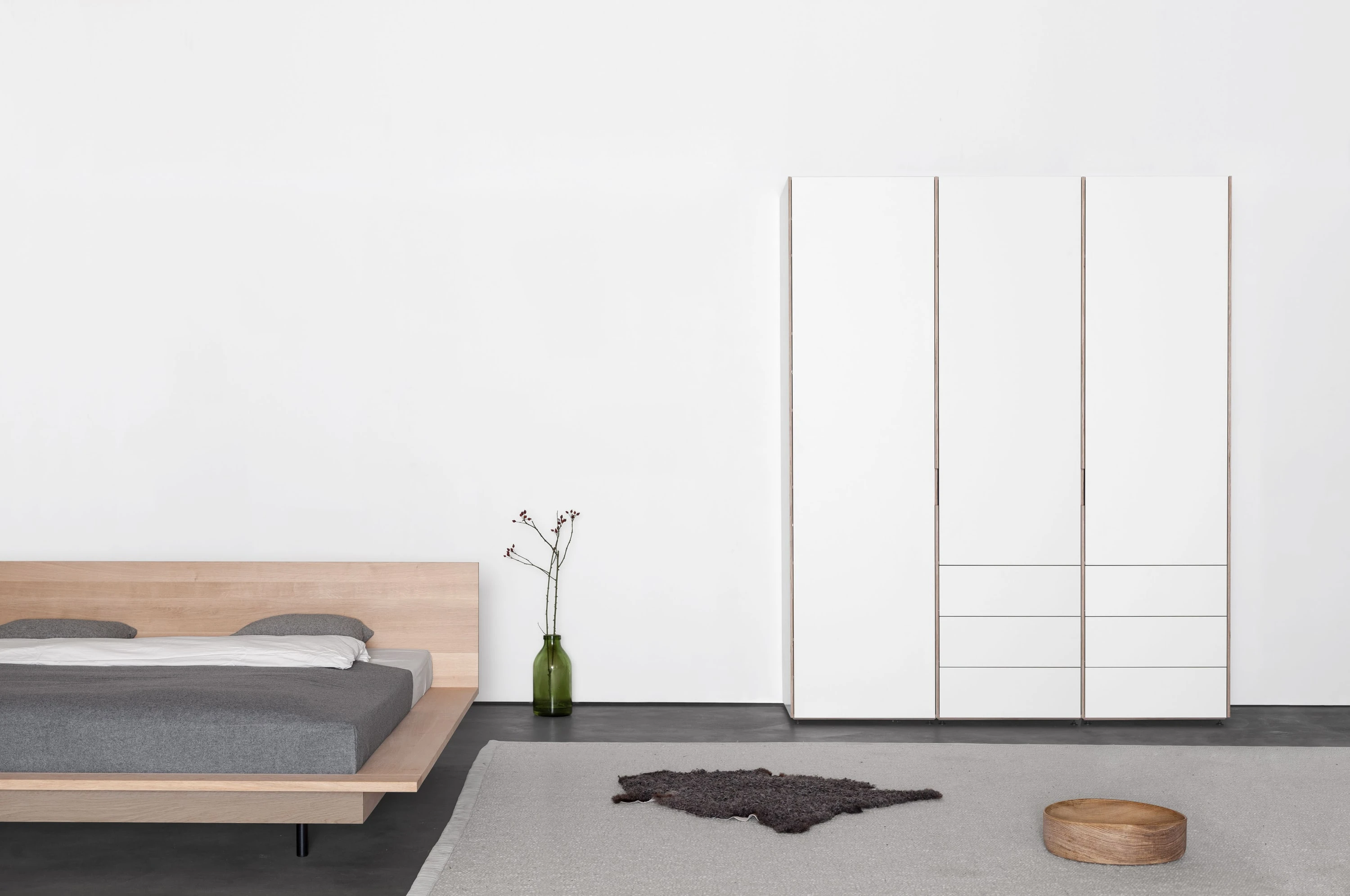 Bett aus Eiche mit grauer Decke vor einem weissen Kleiderschrank mit drei Türen.