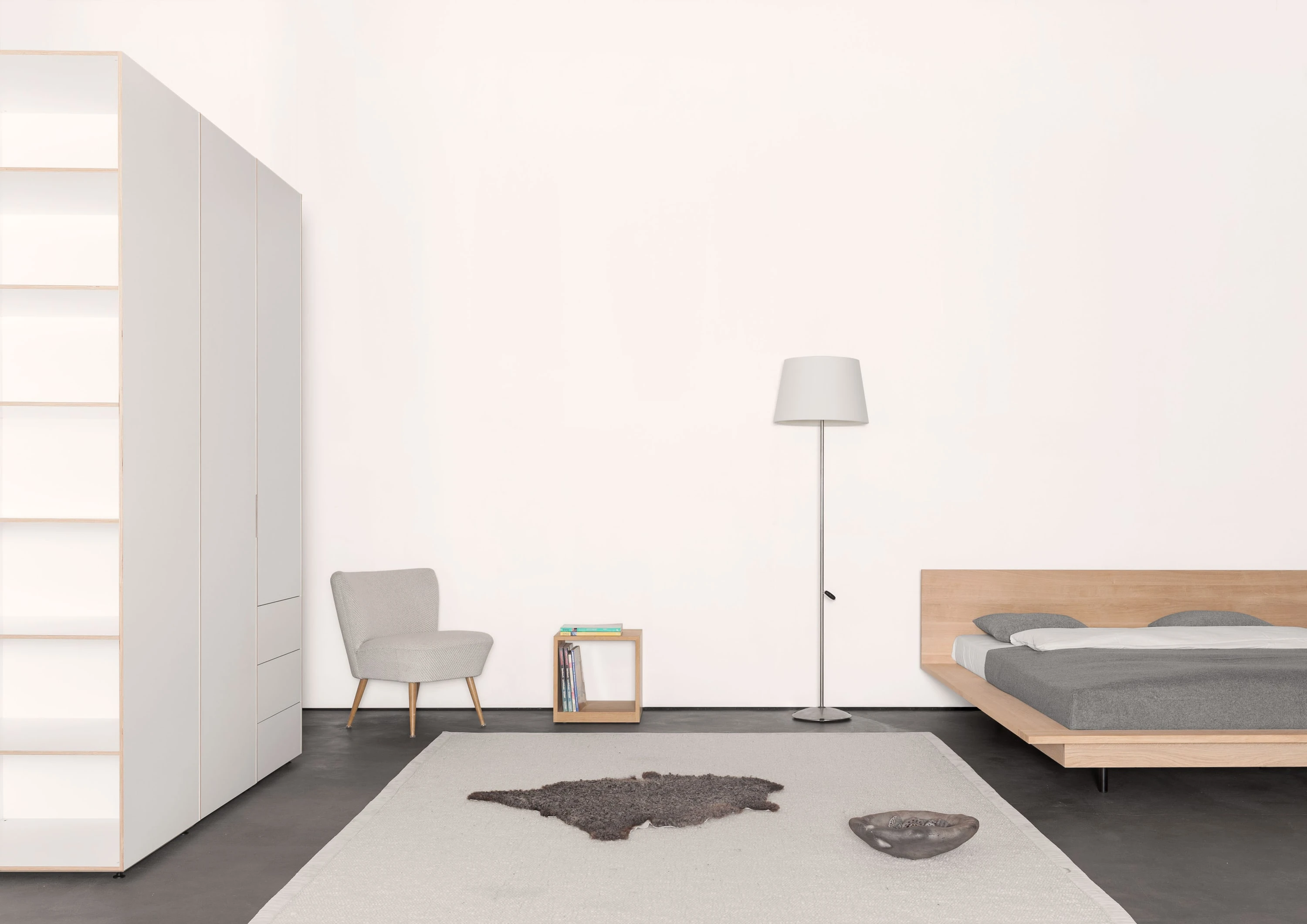 Schlafzimmer mit weissem Kleiderschrank, Sessel, Stehleuchte, Holzkubus und Bett aus Eiche.