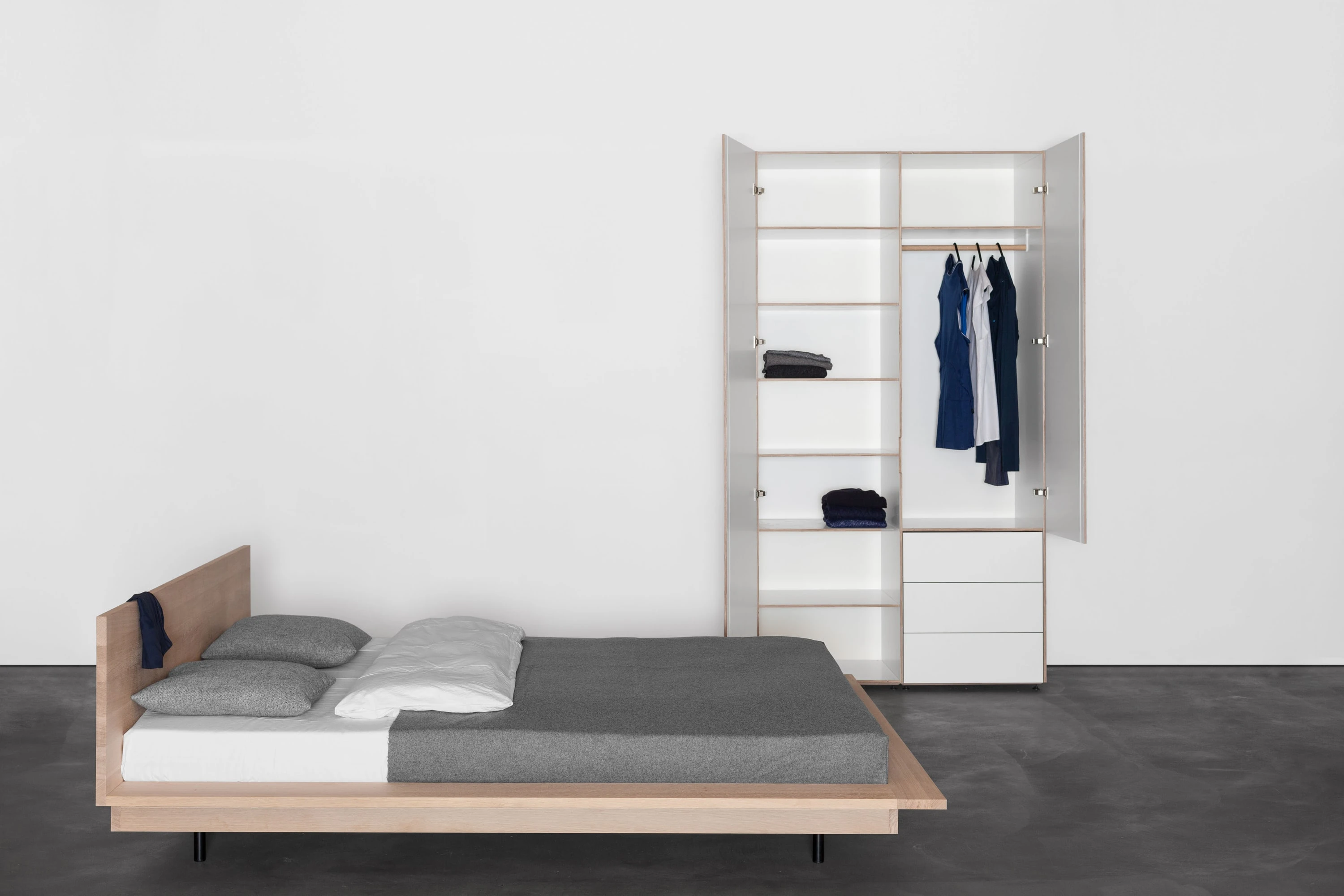 Bett aus Eiche mit grauer Decke, dahinter ein geöffneter weisser Kleiderschrank mit Kleidung.