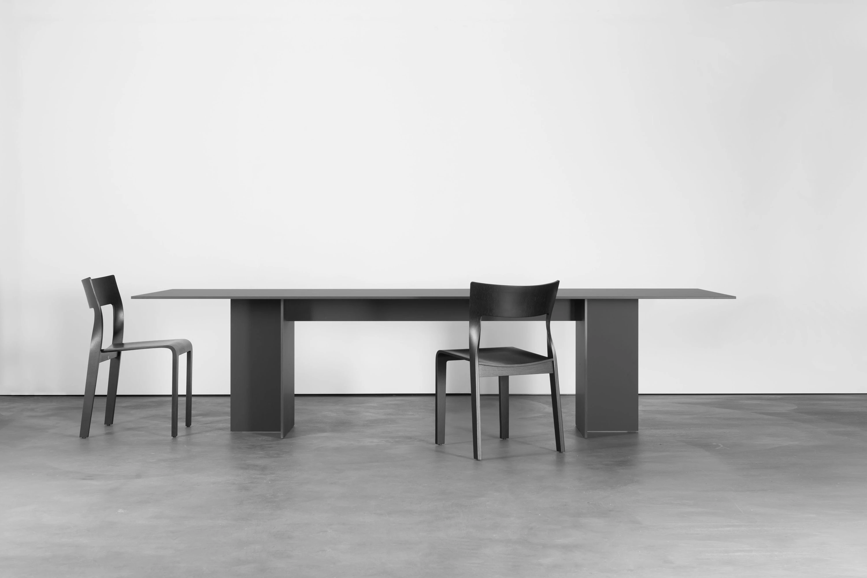 Langer schwarzer Tisch mit zwei schwarzen Stühlen auf hellem Betonboden.