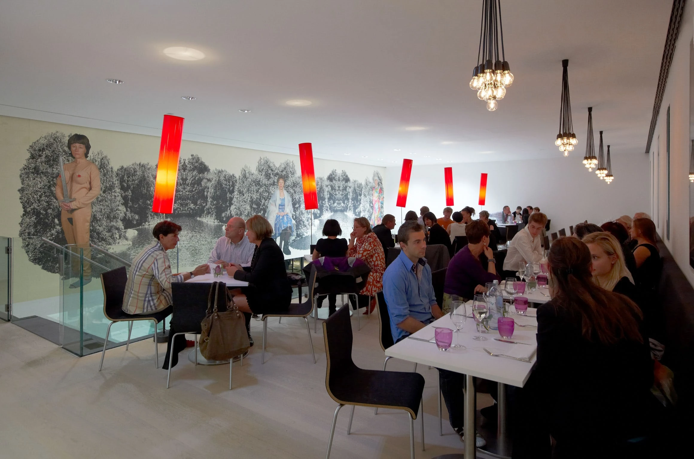 Gut besetztes Restaurant mit weissen Tischen, dunklen Stühlen und roten Leuchten vor einer Wandmalerei.