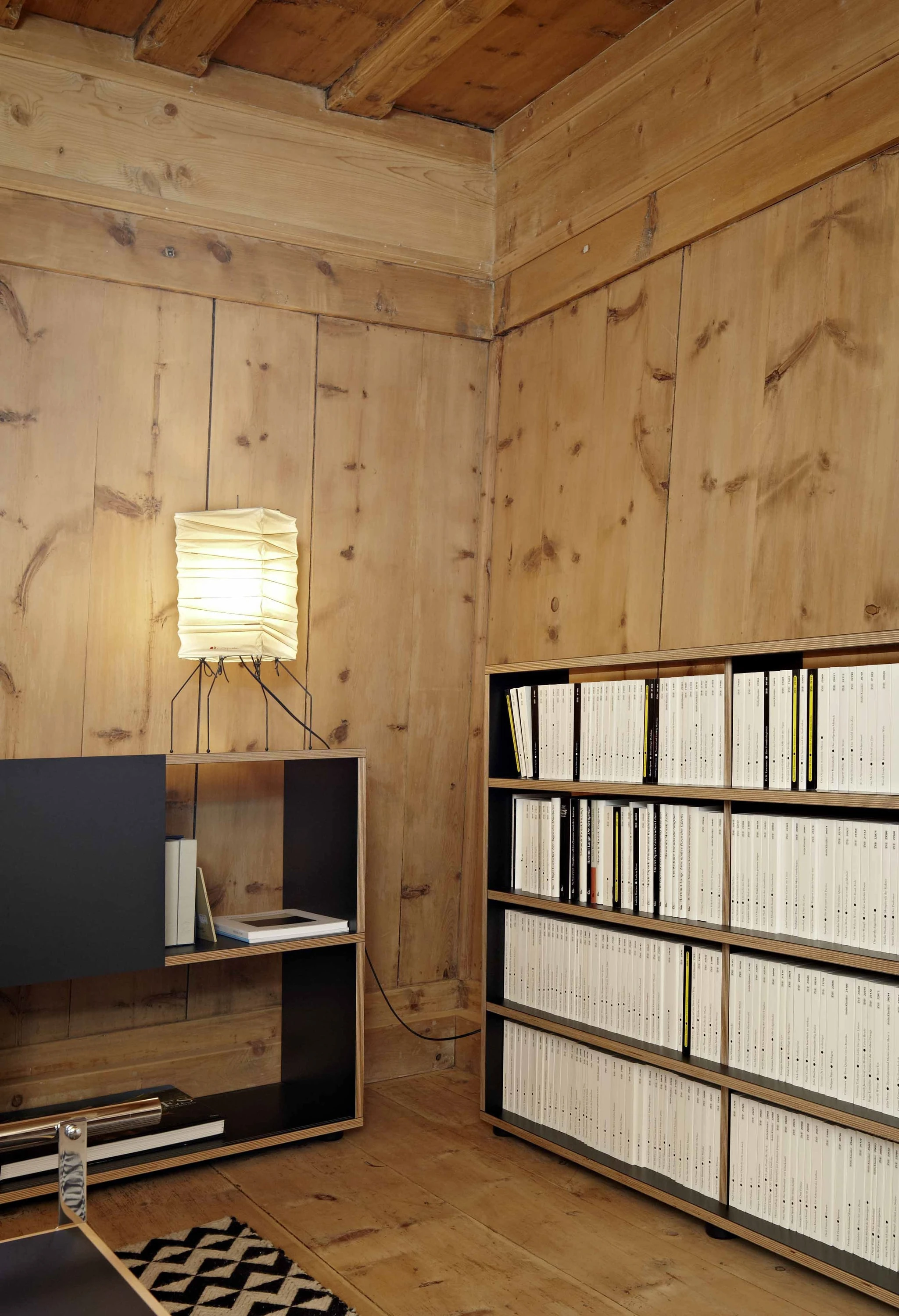 Ecke eines holzvertäfelten Raums mit Leuchte, schwarzem Sideboard und Regal mit Ordnern.