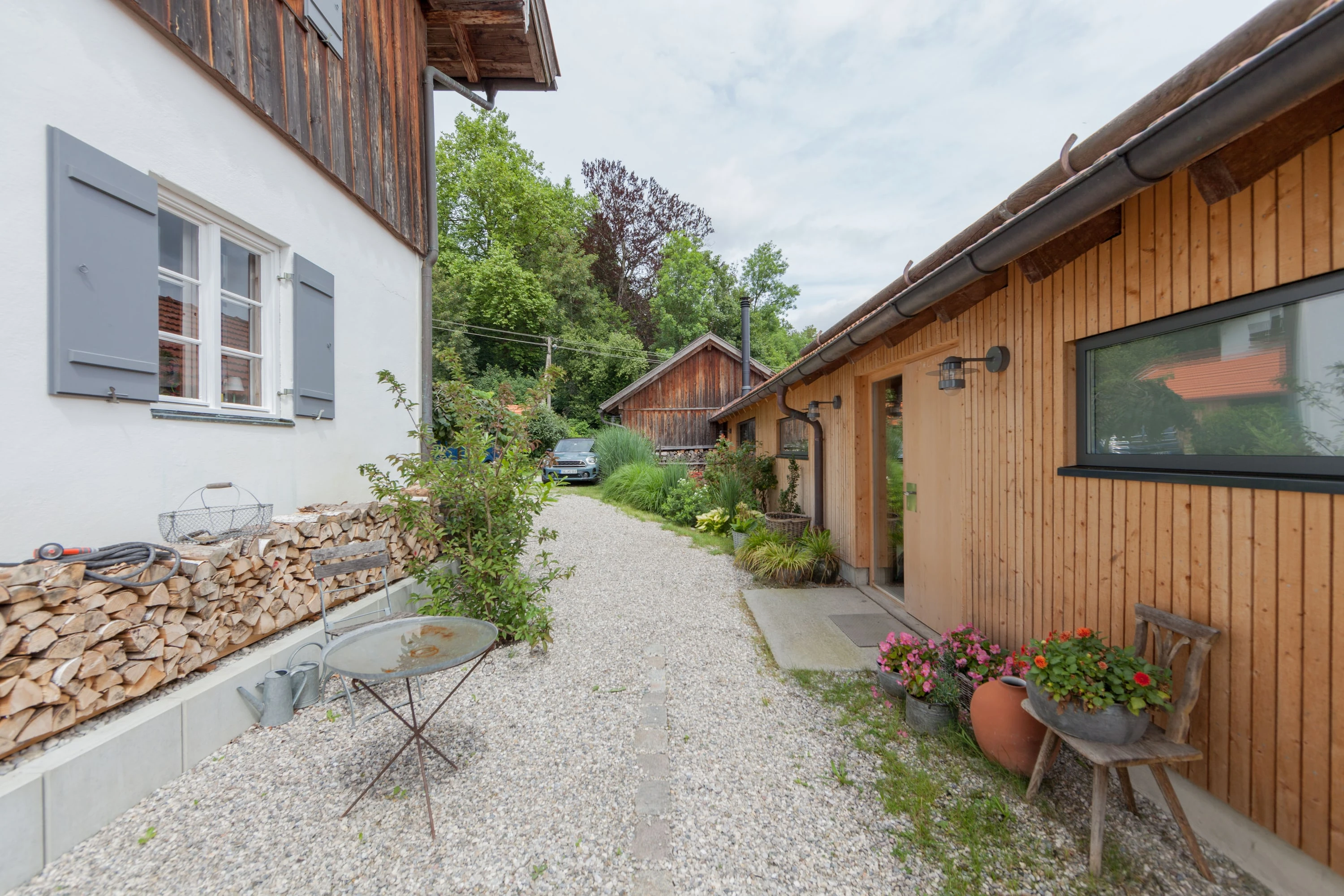 Kiesweg zwischen Bauernhaus und langem Holzgebäude mit Blumen und Brennholz.