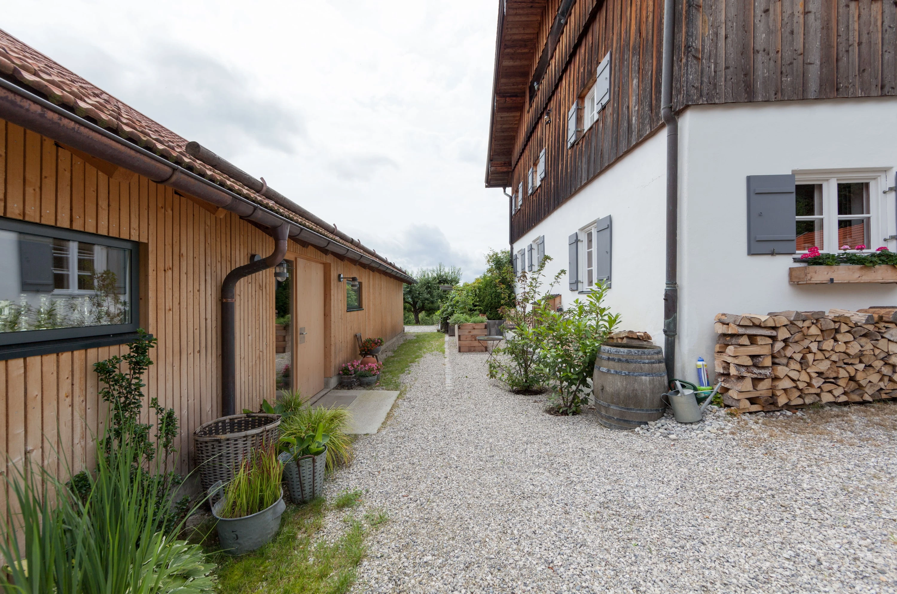 Kiesweg entlang des Holzgebäudes mit Pflanzen und Blick zum Bauernhaus.