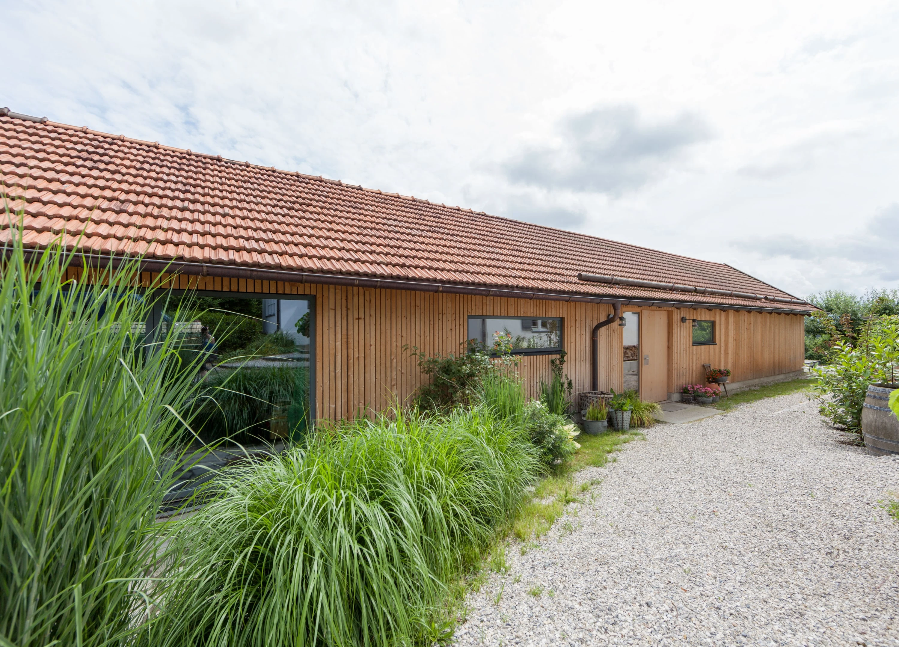Langgestrecktes Holzgebäude mit Ziegeldach hinter hohen Gräsern.