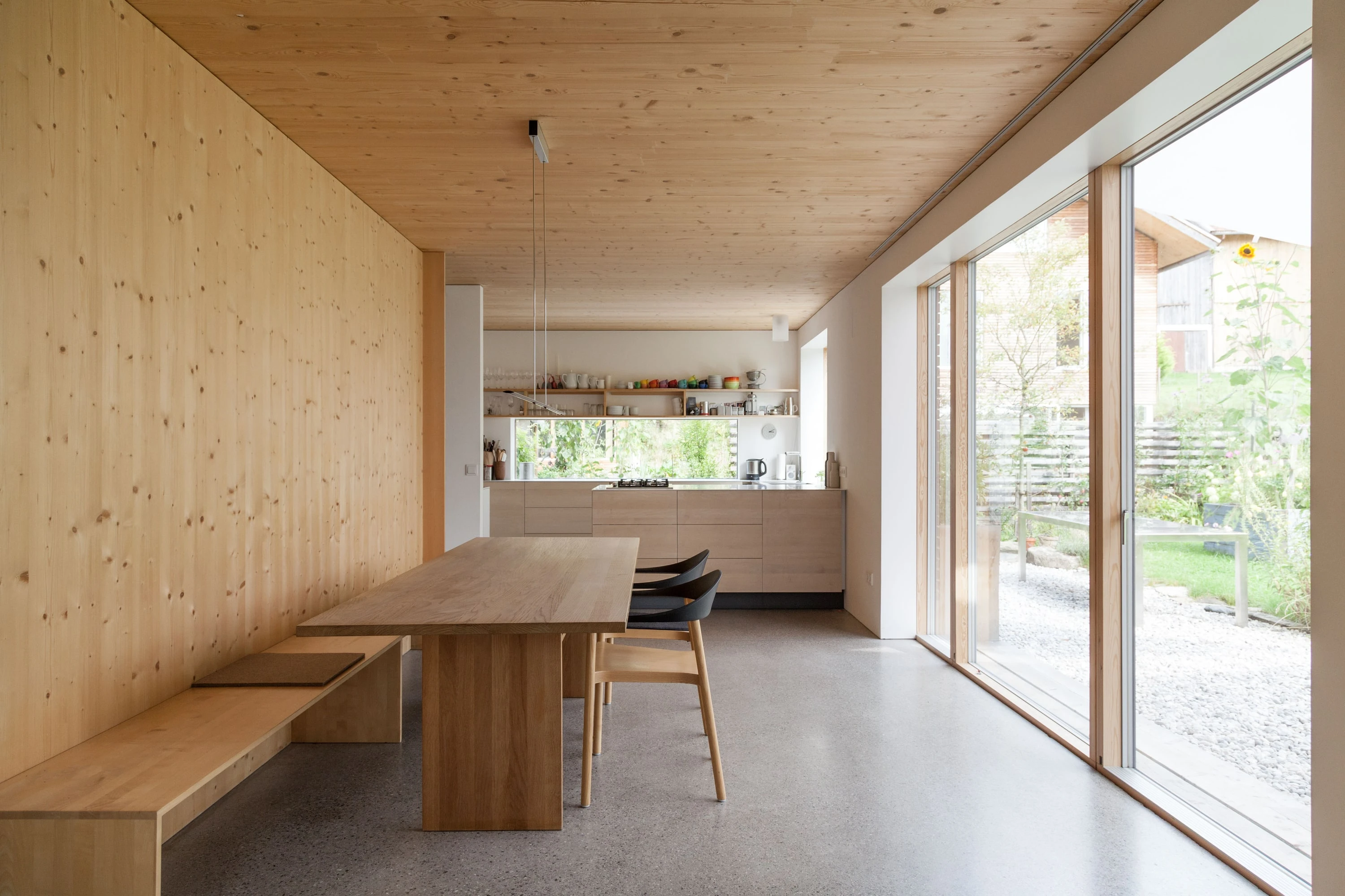 Essraum mit Holzwänden, langem Holztisch mit Bank und grosser Glasfront zum Garten.