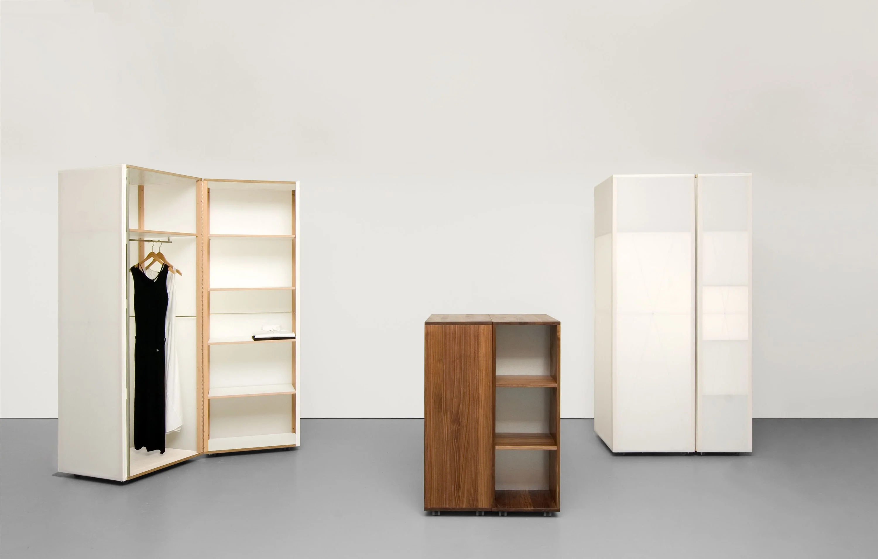 Geöffneter Rollschrank mit Textilbespannung und Kleid, daneben ein kleiner Schrank aus dunklem Holz und ein geschlossener weisser Schrank.
