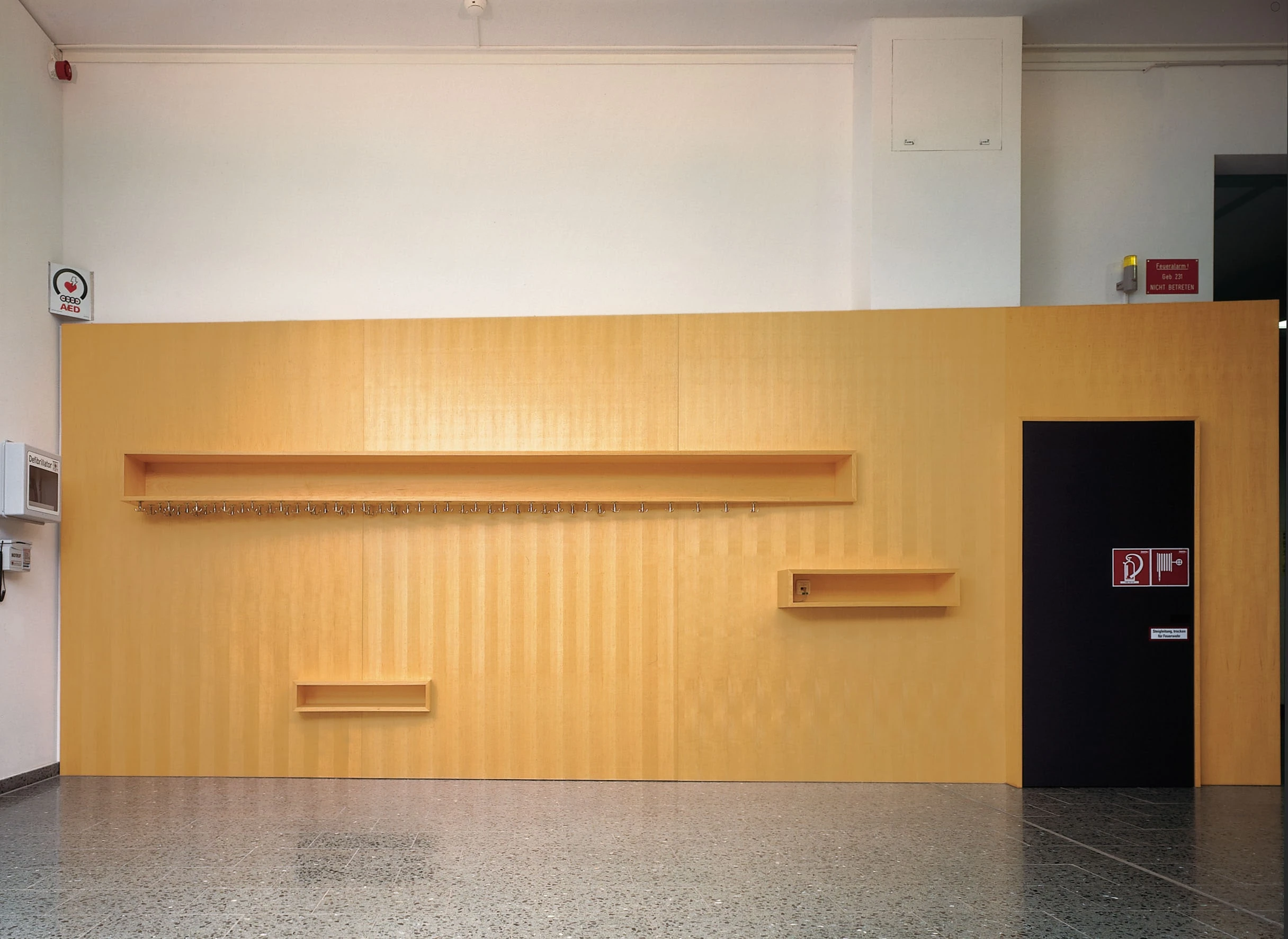 Holzwand mit Ablagen und Garderobe neben einer schwarzen Tür im Foyer.